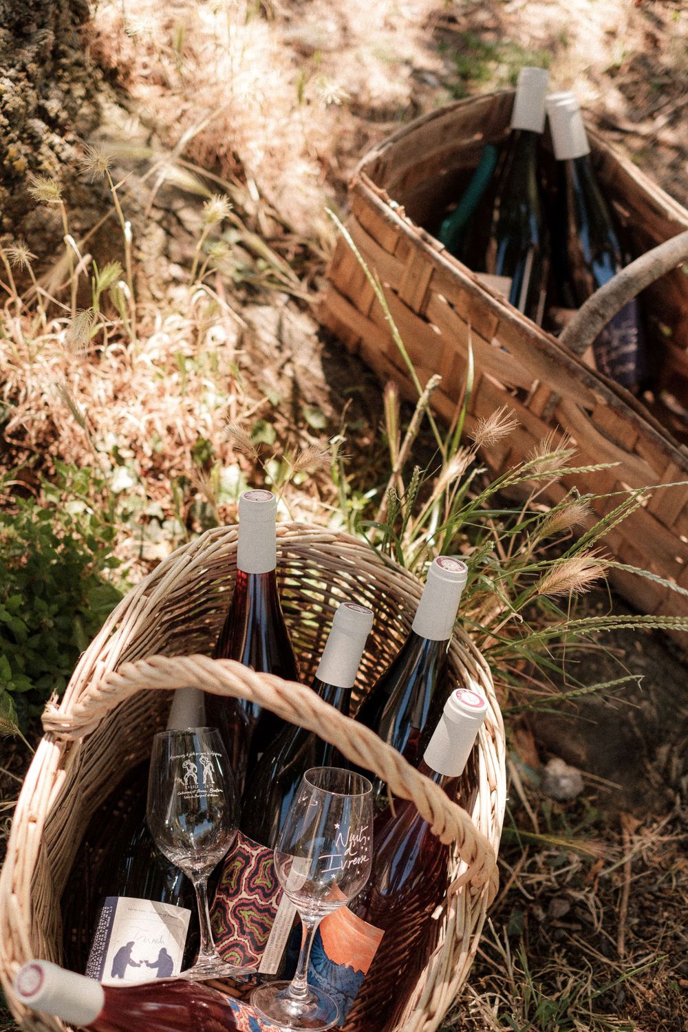 Wine bottles and glasses arranged in rustic woven baskets among dried grass.