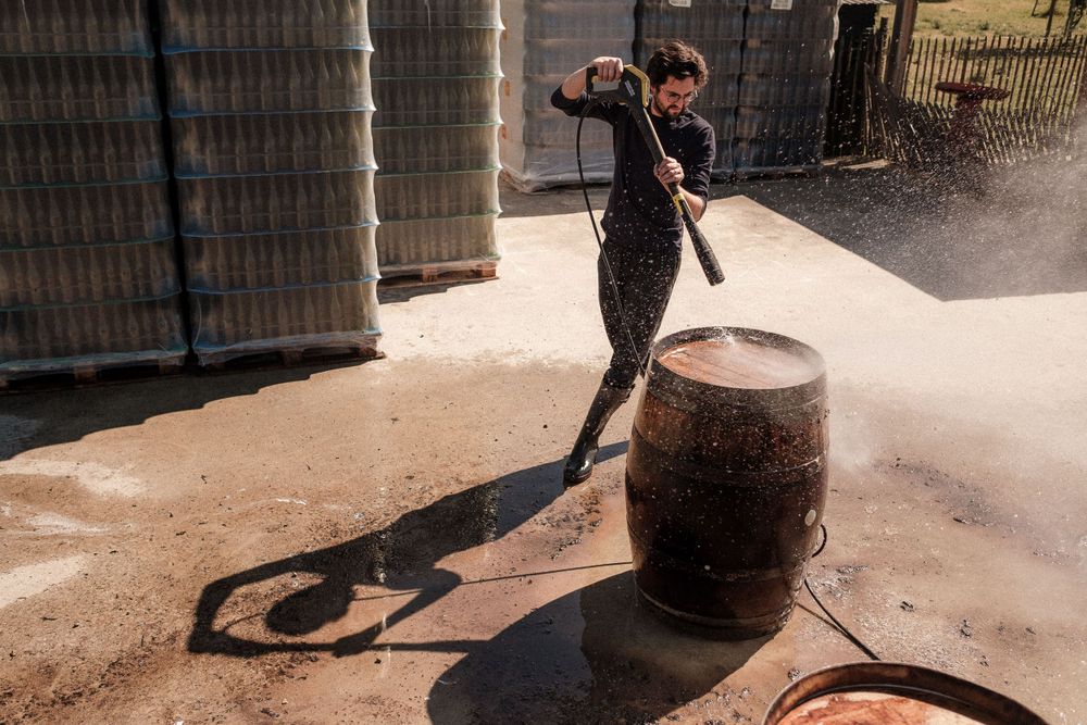 A person cleaning or maintaining a wooden wine barrel in a warehouse setting, creating a dramatic shadow on the floor.