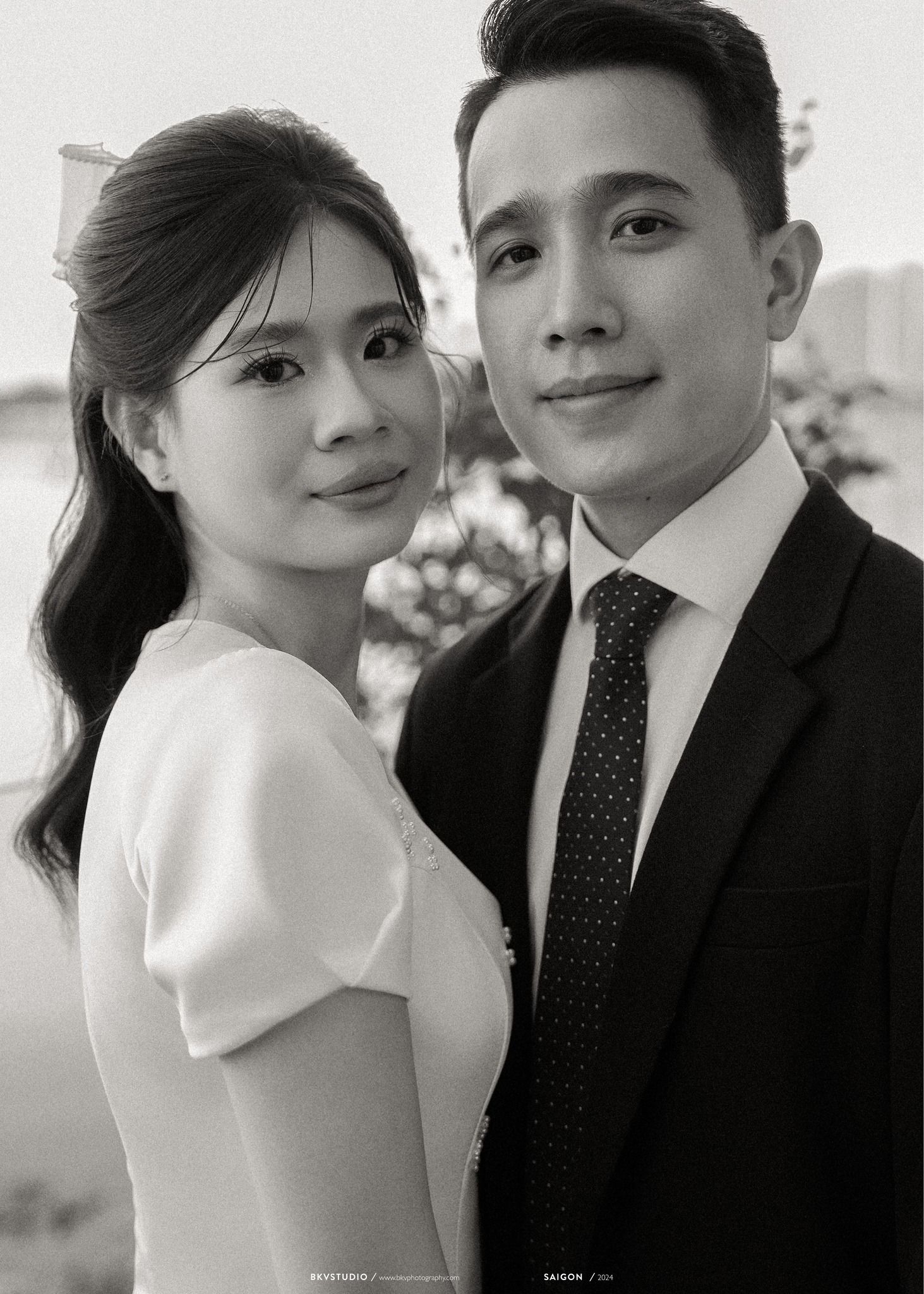 A black and white portrait of a couple in formal attire, with her in a white dress and him in a dark suit and polka dot tie.