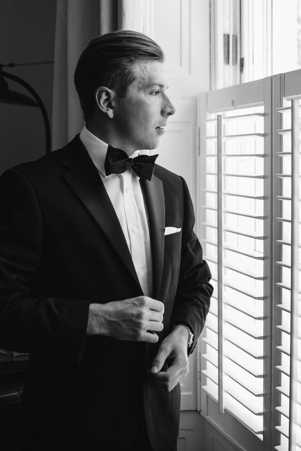 A black and white photograph shows someone in a classic tuxedo with bow tie looking pensively through window blinds at The Halifax Club