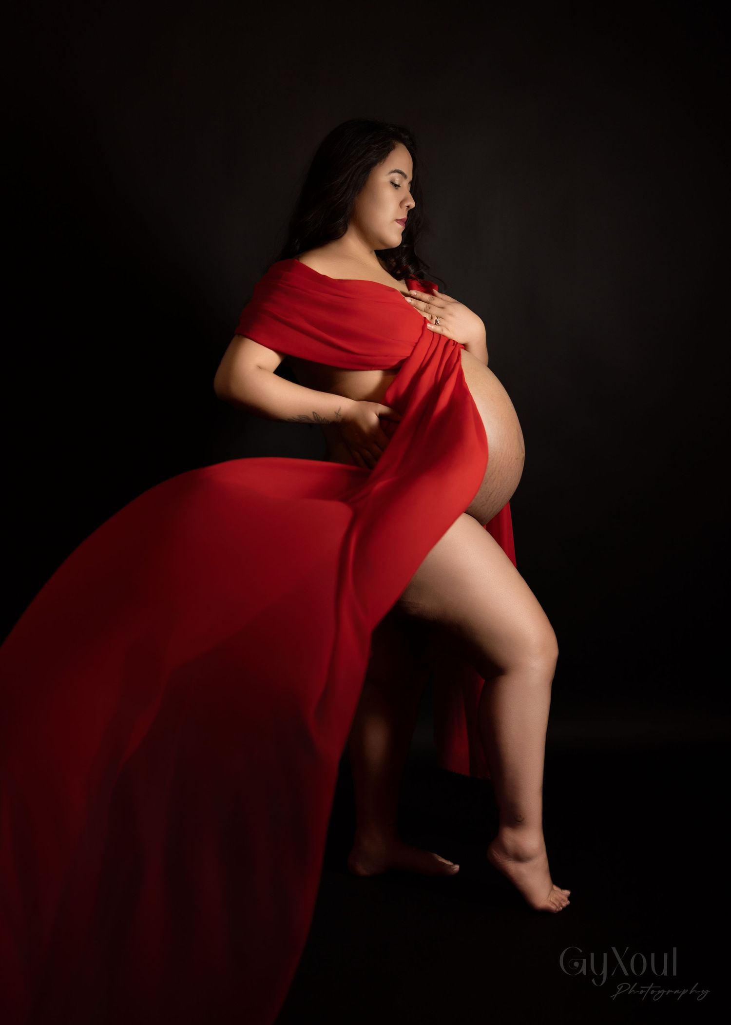 Dramatic maternity portrait with billowing red fabric in a dark studio setting.