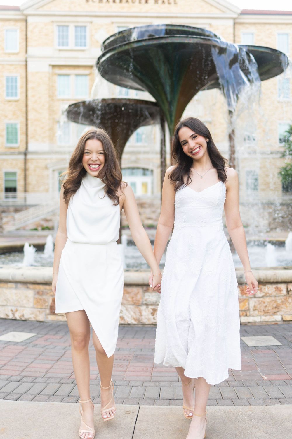 Two young women in white dresses hold hands and smile in front of a decorative fountain outside Scharbauer Hall.