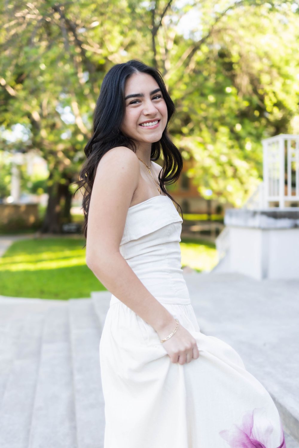 Young woman in white strapless dress smiling outdoors in lush green garden setting.