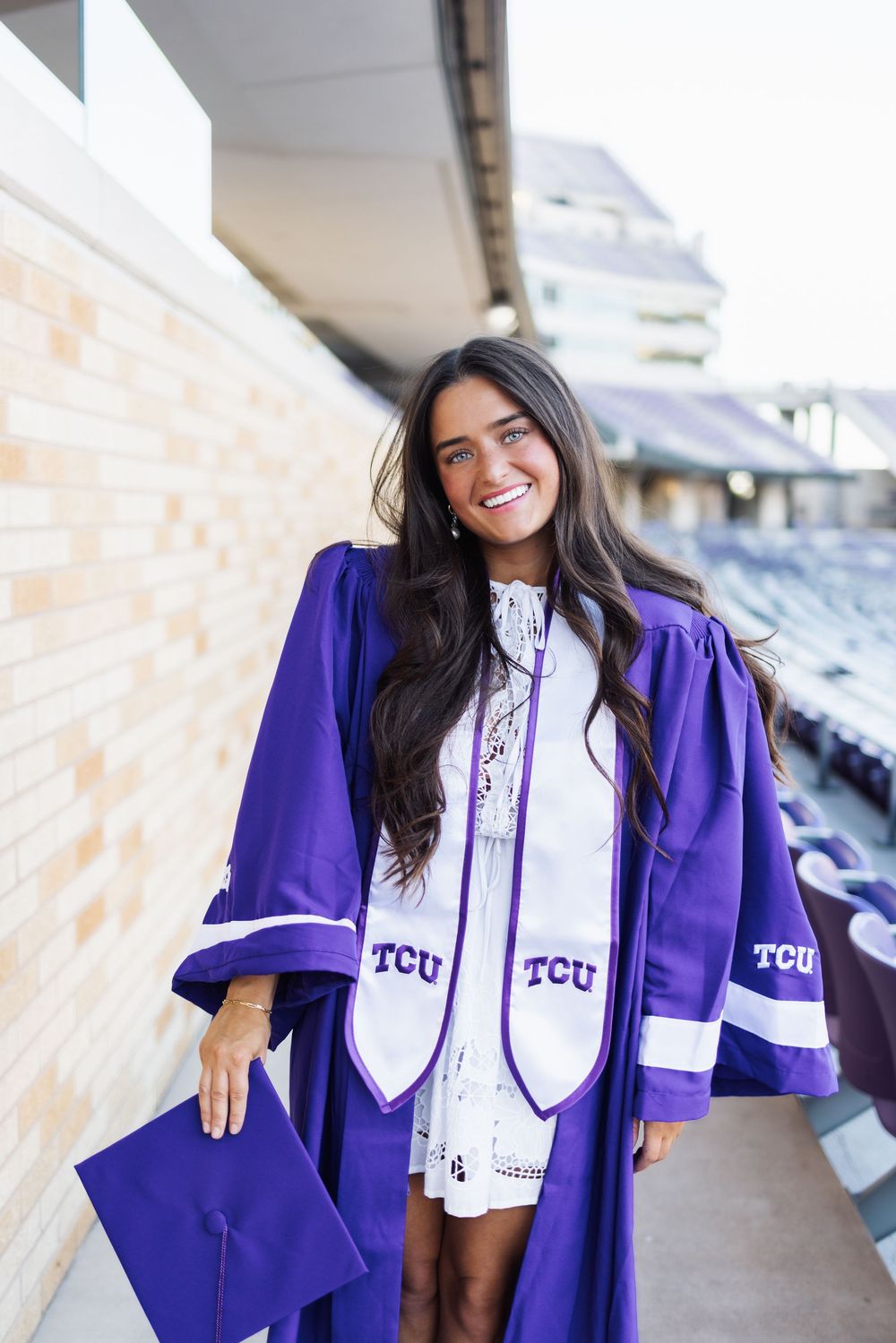 Graduate wearing purple TCU graduation gown and cap smiles while posing outside on campus.