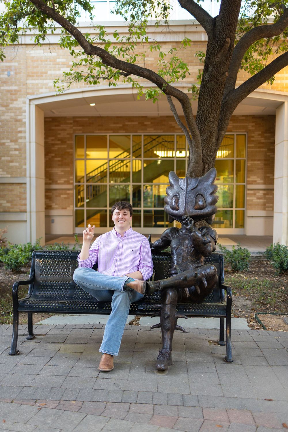 Man in pink shirt sitting on bench next to a bronze mascot statue outside a brick building.