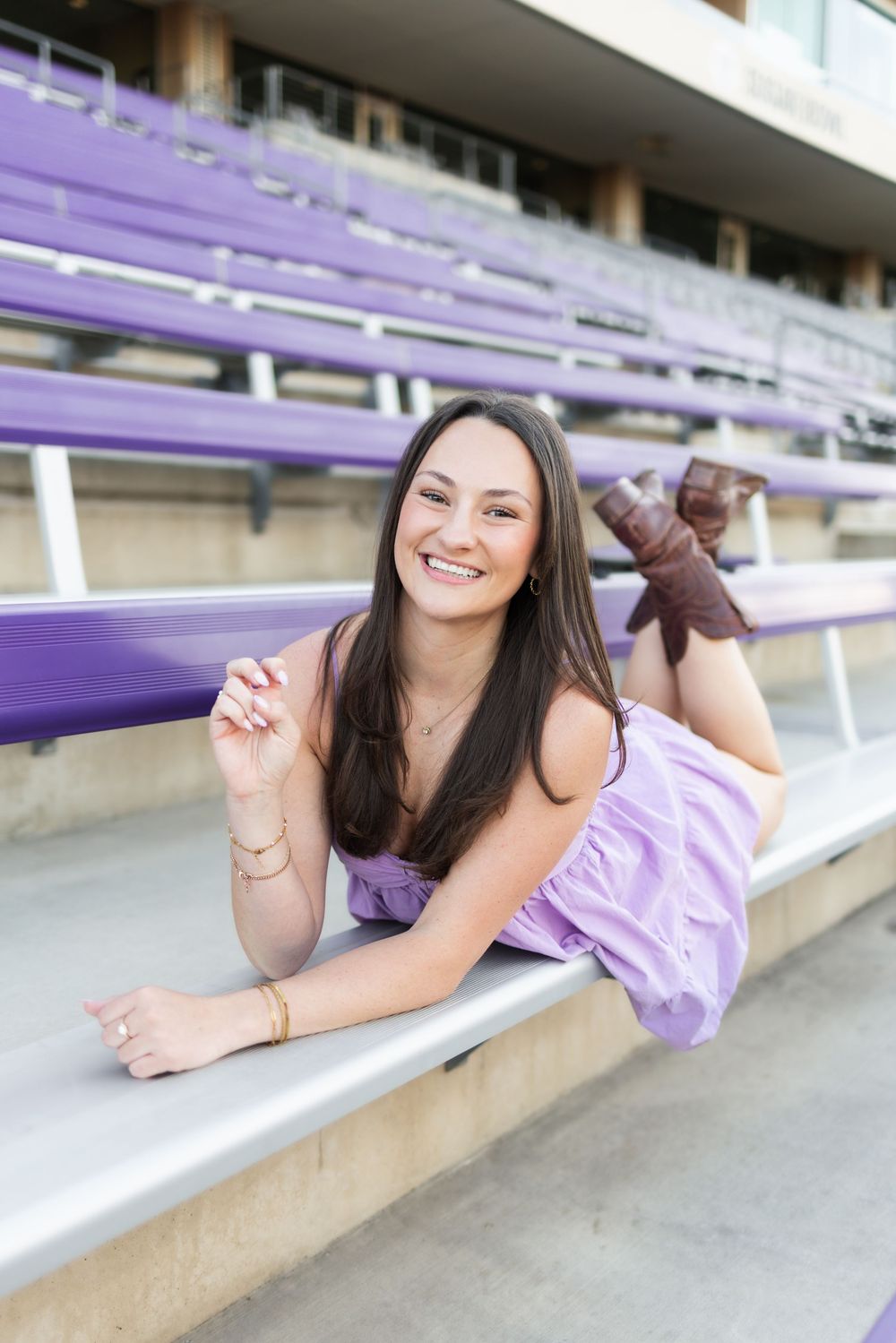 Young woman in lavender dress and brown boots smiling while lying on purple bleachers at a stadium.