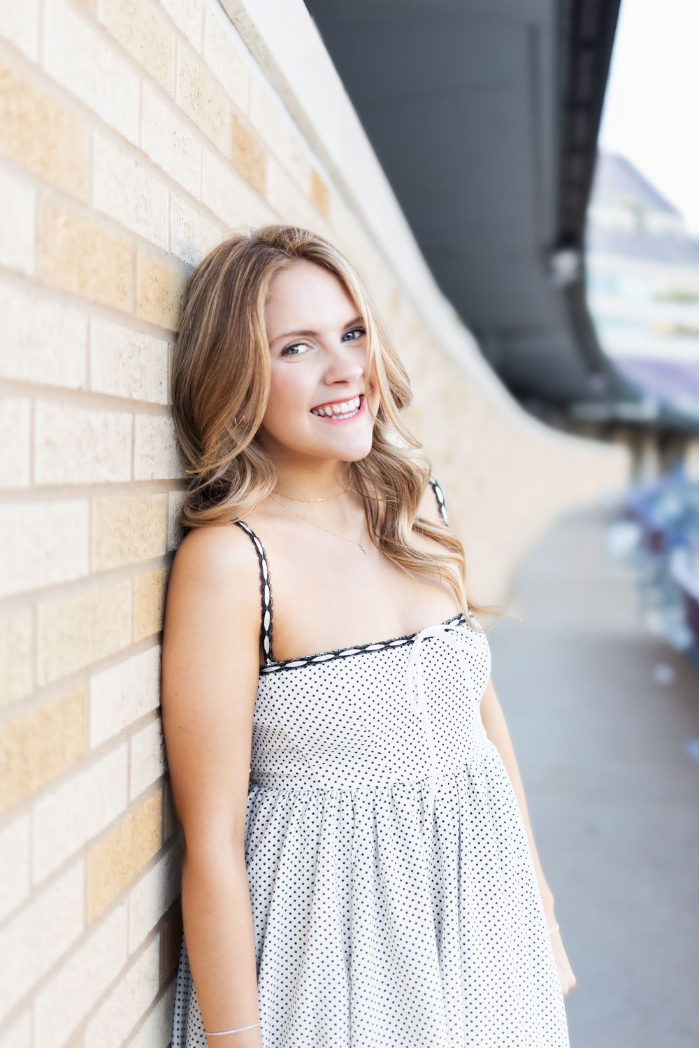 Young woman in white polka dot sundress smiling and leaning against a brick wall outdoors.