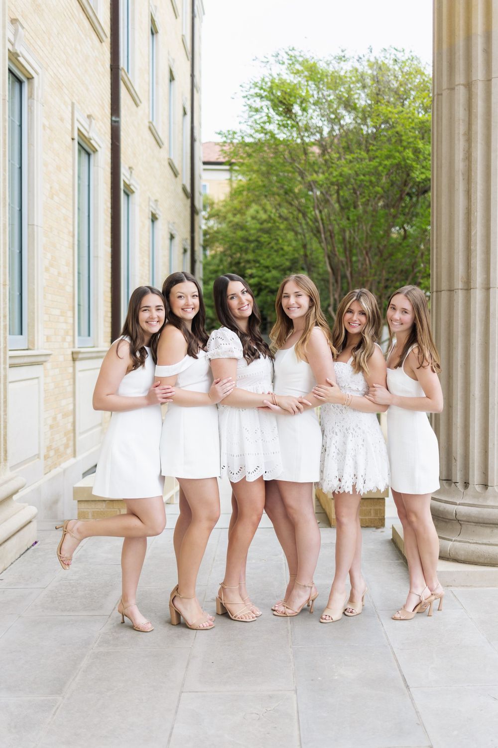 Six young women in matching white dresses and nude heels pose together outdoors near a classic stone building.