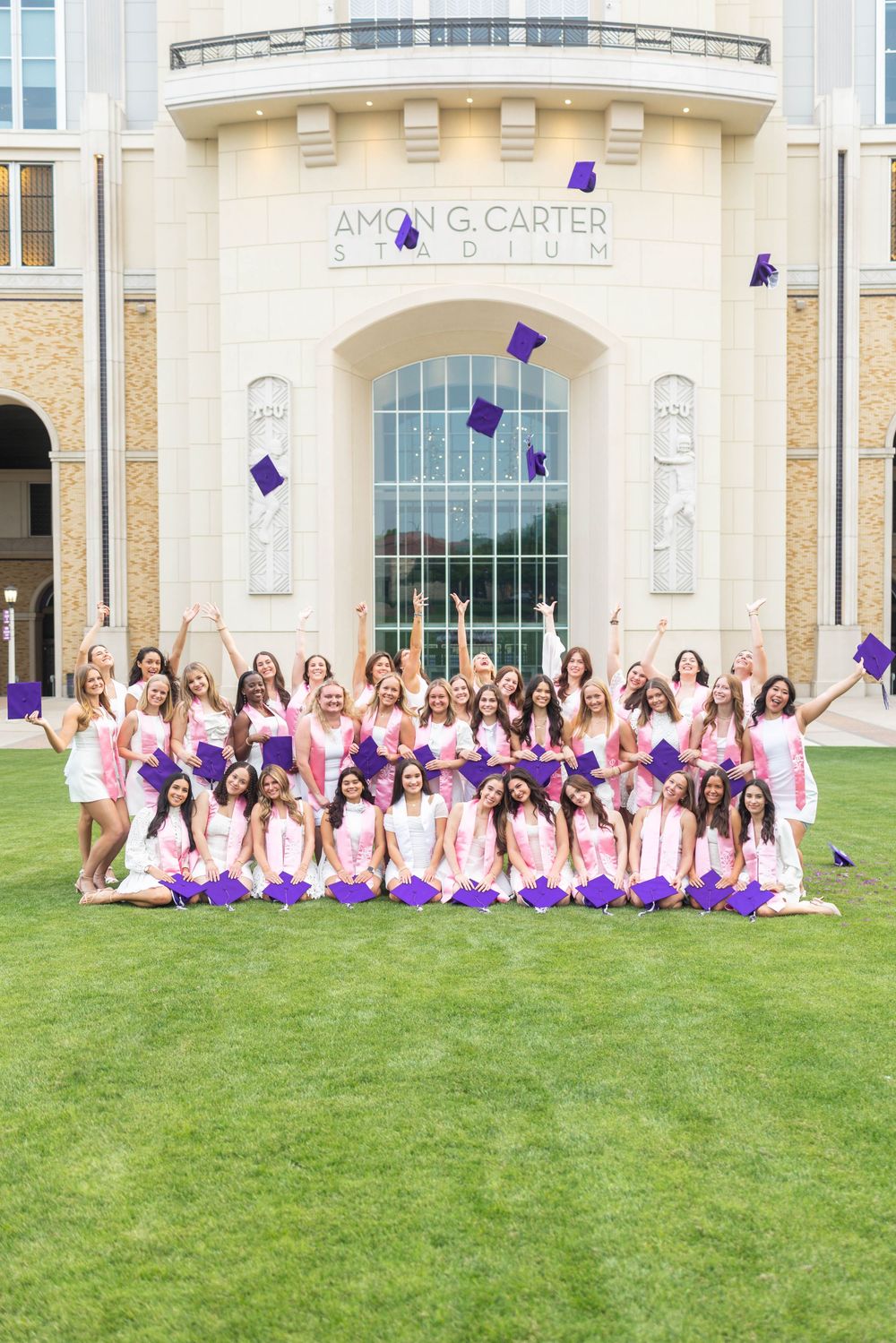 Group of graduating women in pink dresses tossing purple caps at Amon G. Carter Stadium celebration.