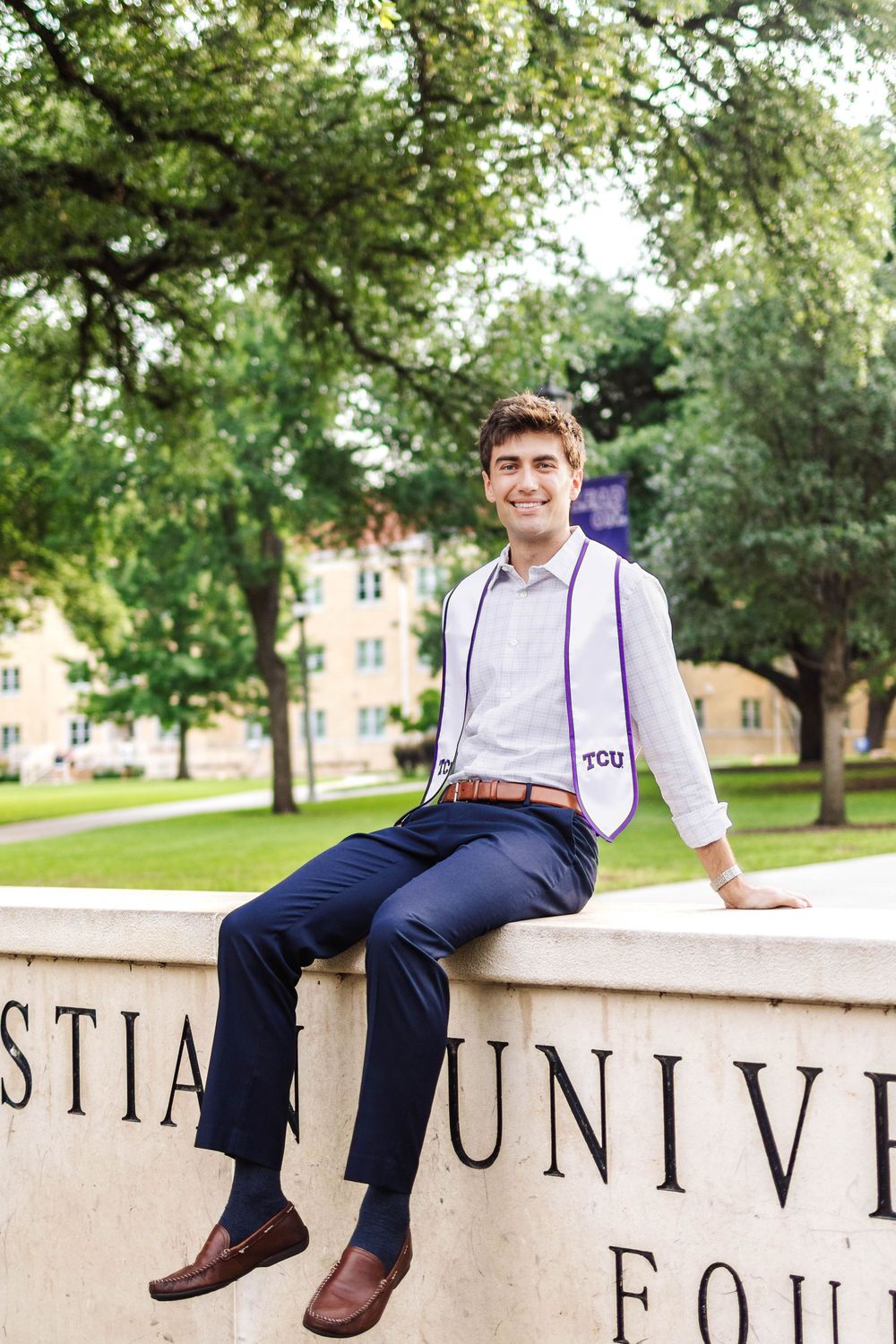 A person in a light shirt and suspenders sits casually on a stone wall marked with 'STIAN UNIV' text in an outdoor setting.