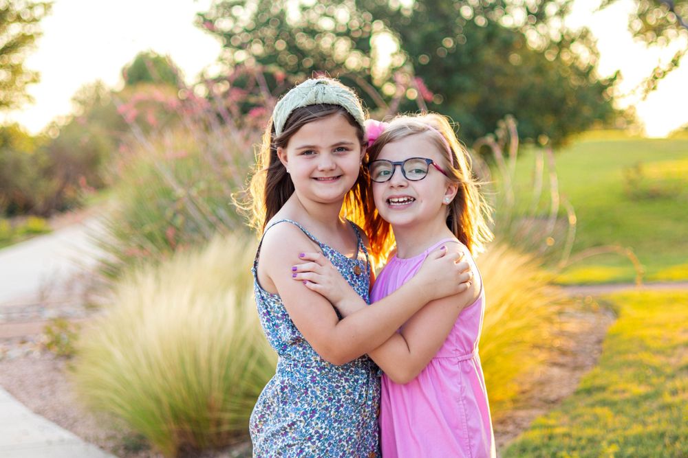 Two children in colorful dresses embrace and smile during sunset in a garden setting.