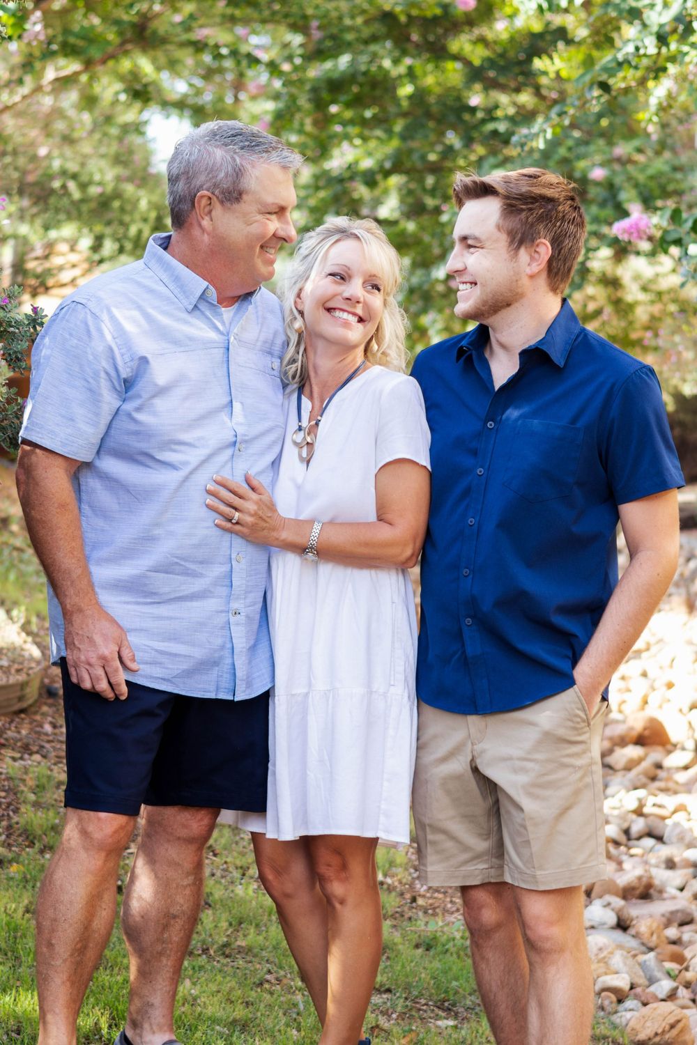 Three people share a candid laugh together while posing for a family photo in a garden setting.
