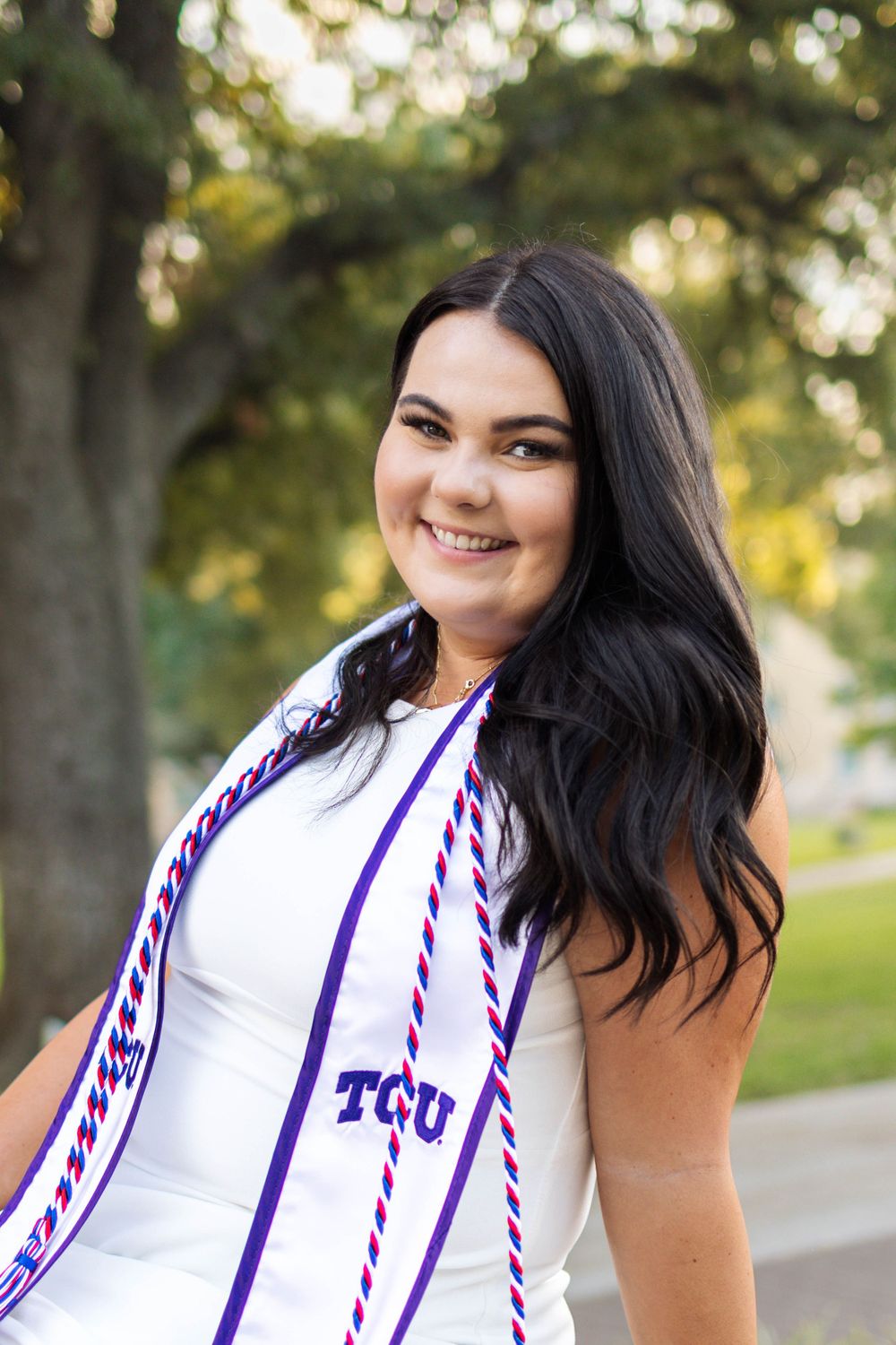 Graduate wearing white dress and purple TCU graduation stole standing outdoors on campus with greenery in background.