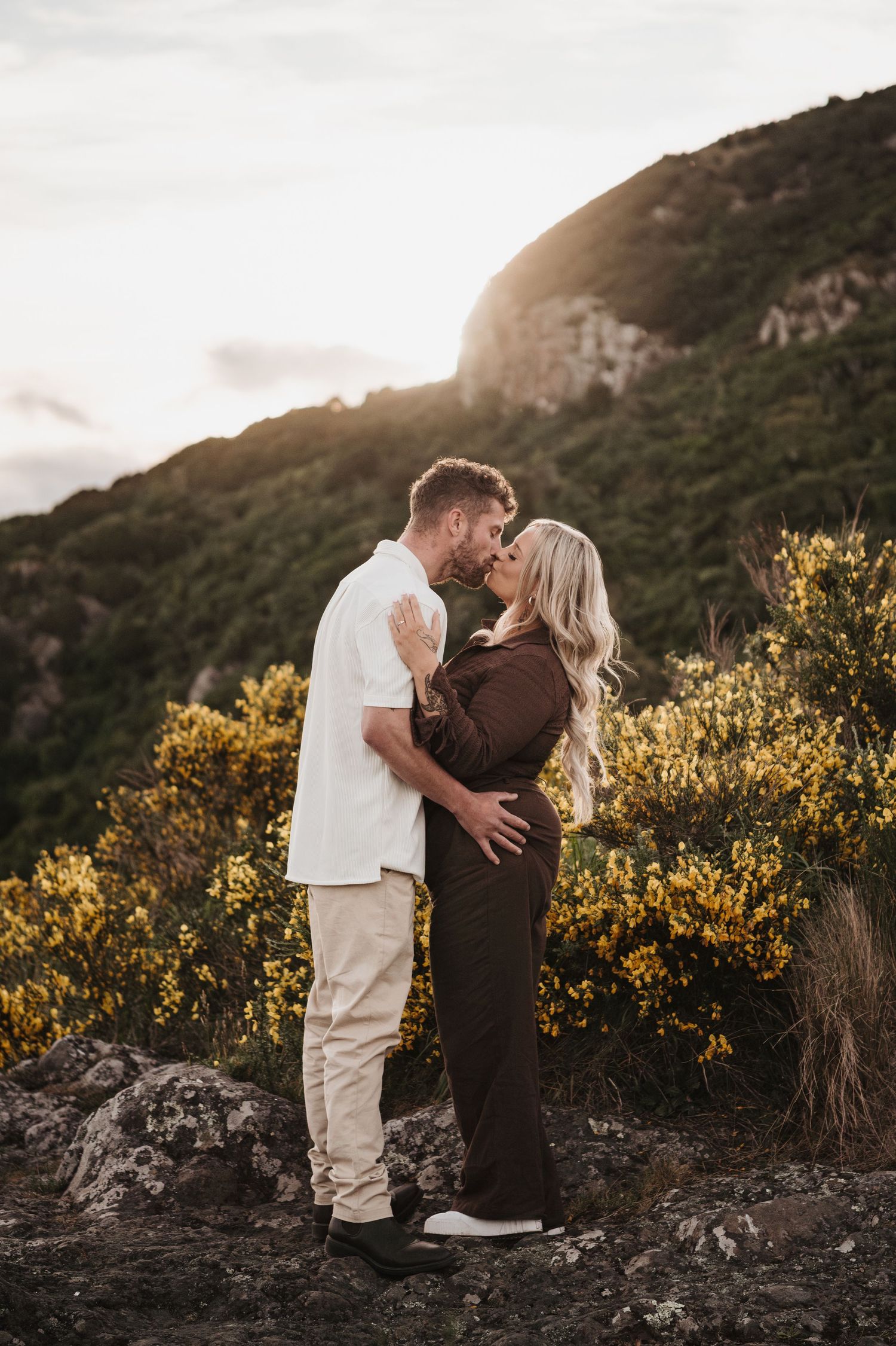 A couple shares an intimate moment against a sunset mountain backdrop with yellow wildflowers.