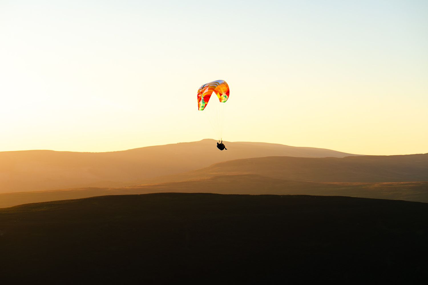Silhouette of orange paraglider soaring over rolling desert dunes at sunset against golden sky.