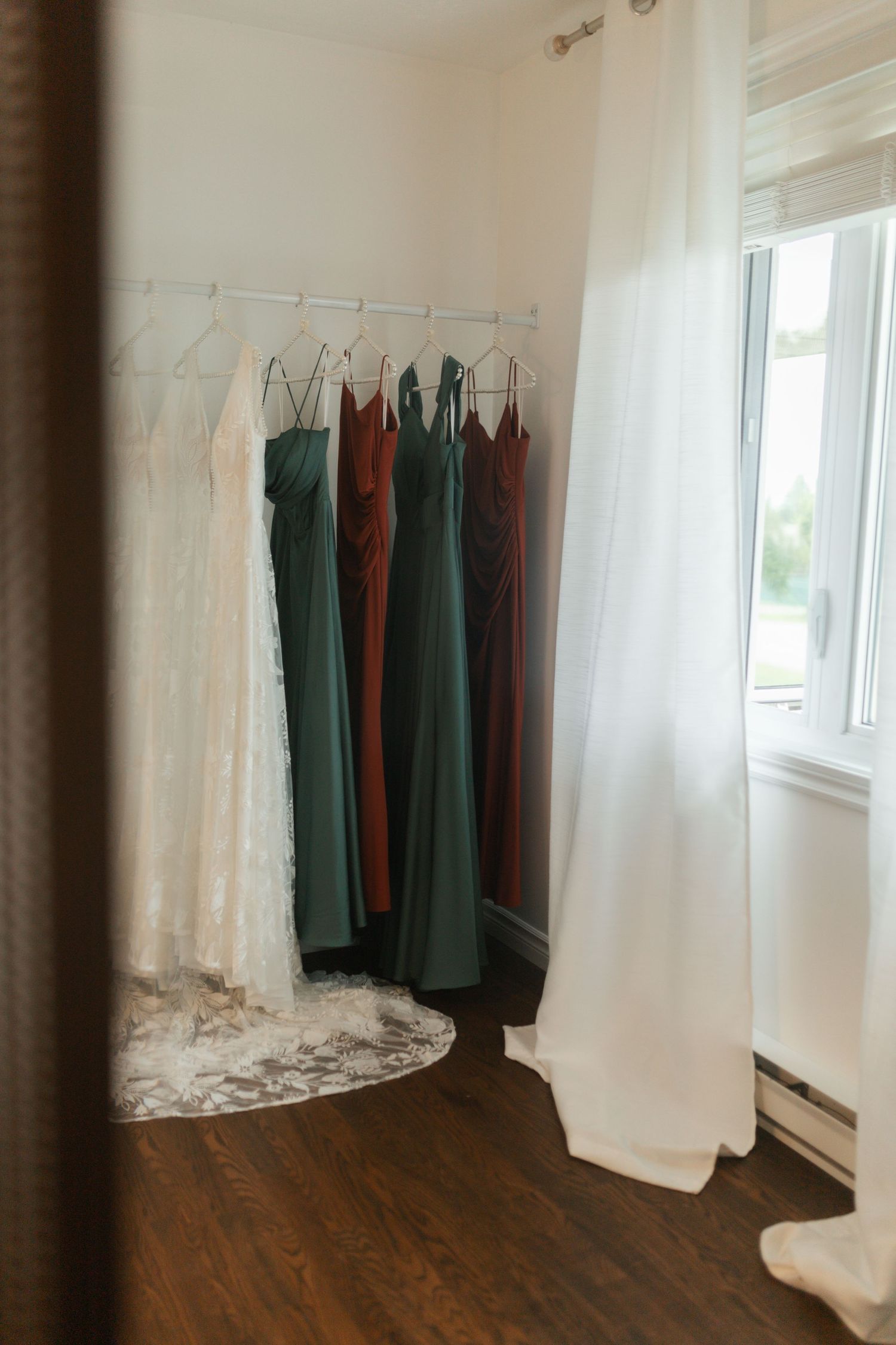 Wedding dresses on a rack including white bridal gown and green and rust bridesmaid dresses near a window.