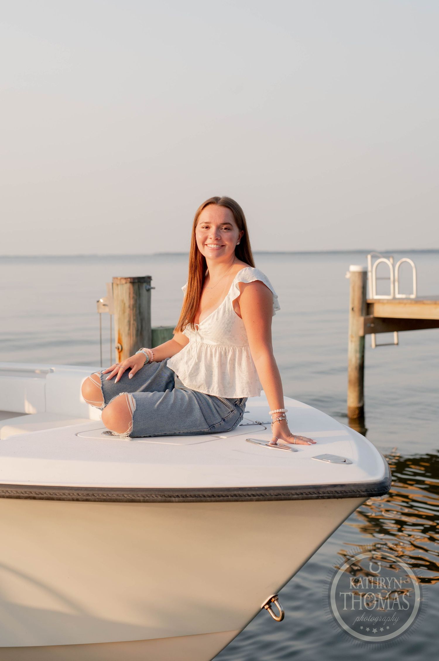 Woman in white top and jeans relaxes on boat deck during golden hour by waterfront dock.