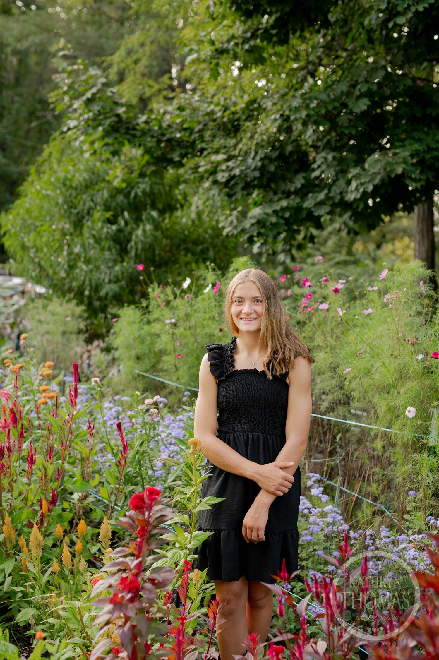 A person in a black dress stands smiling in a garden filled with colorful summer flowers.