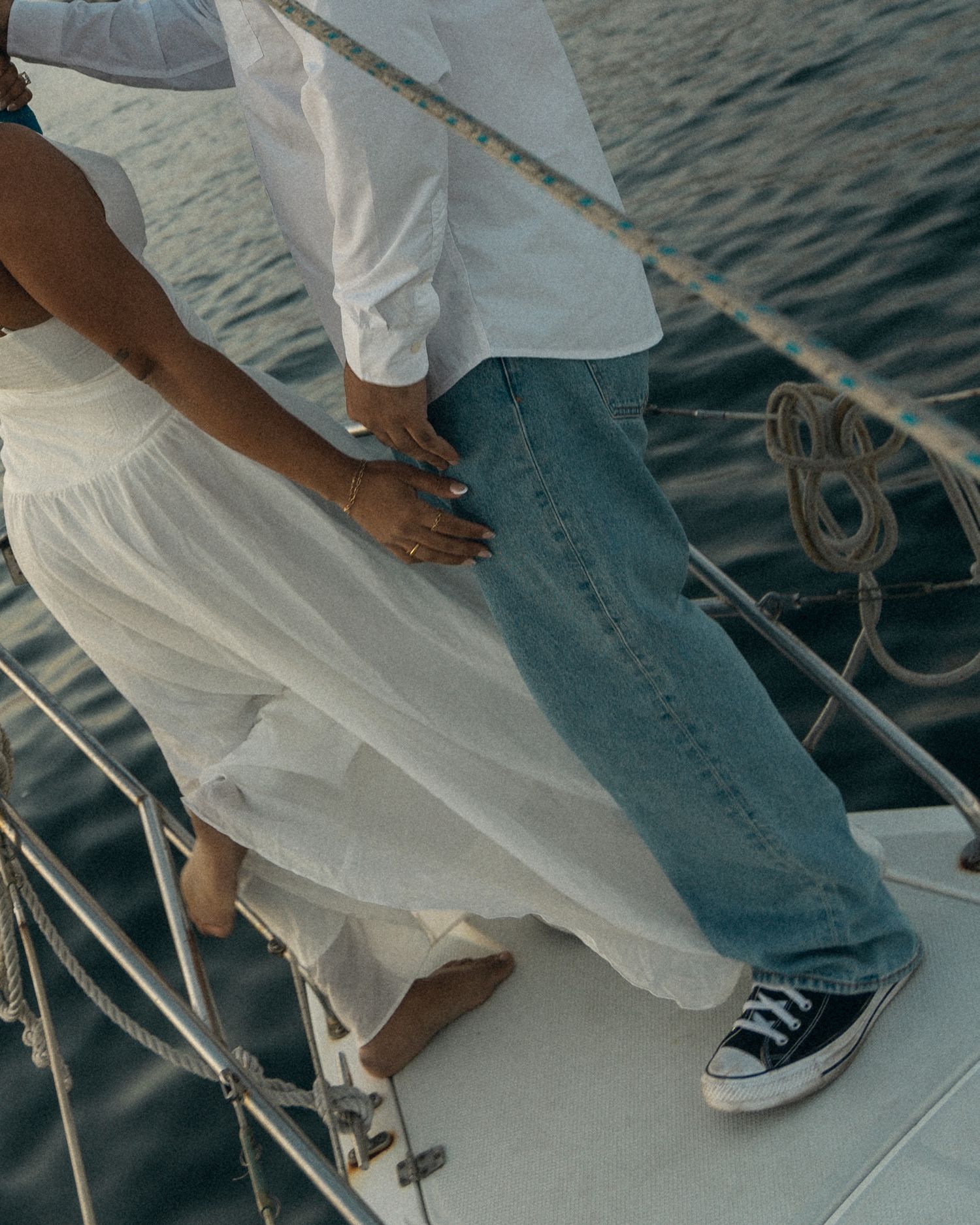 Cropped view of legs in casual attire standing on a sailboat deck with rope coils against blue ocean water.