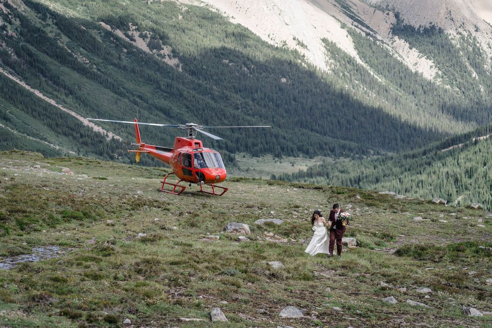 Rockies Heli Wedding A red helicopter lands on a mountain meadow while people pose for a scenic mountain wedding photo shoot.