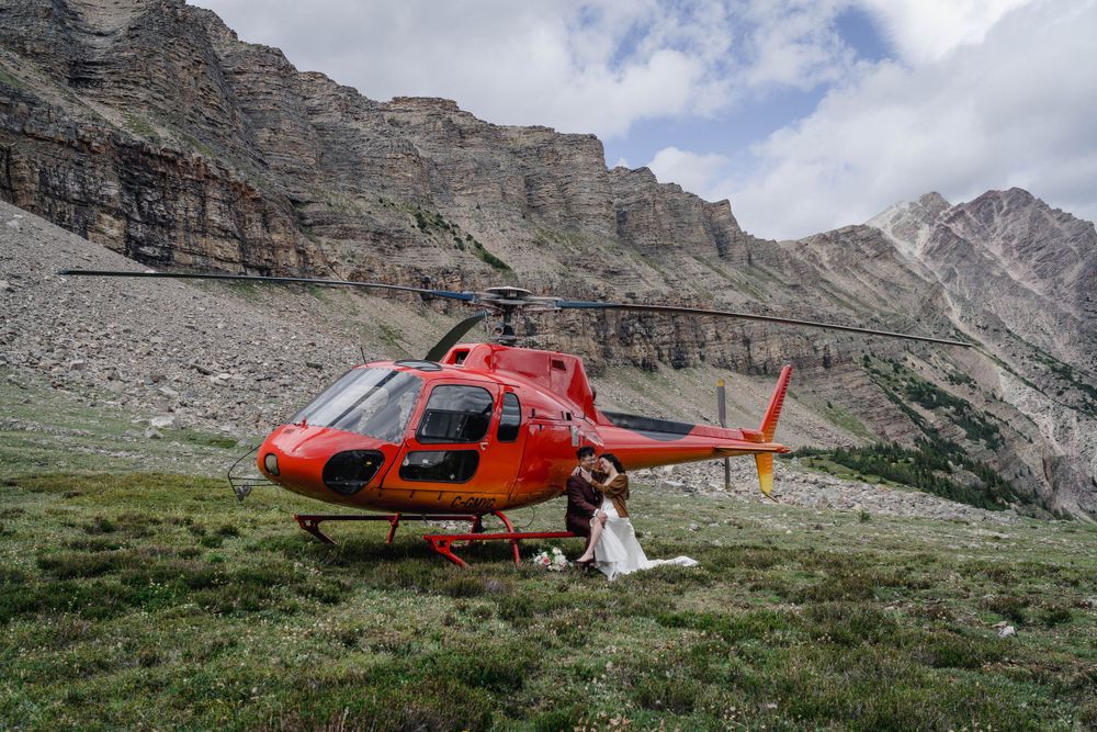 Rockies Heli wedding A red rescue helicopter sits on a grassy mountain slope against towering rocky cliffs in the Dolomites.