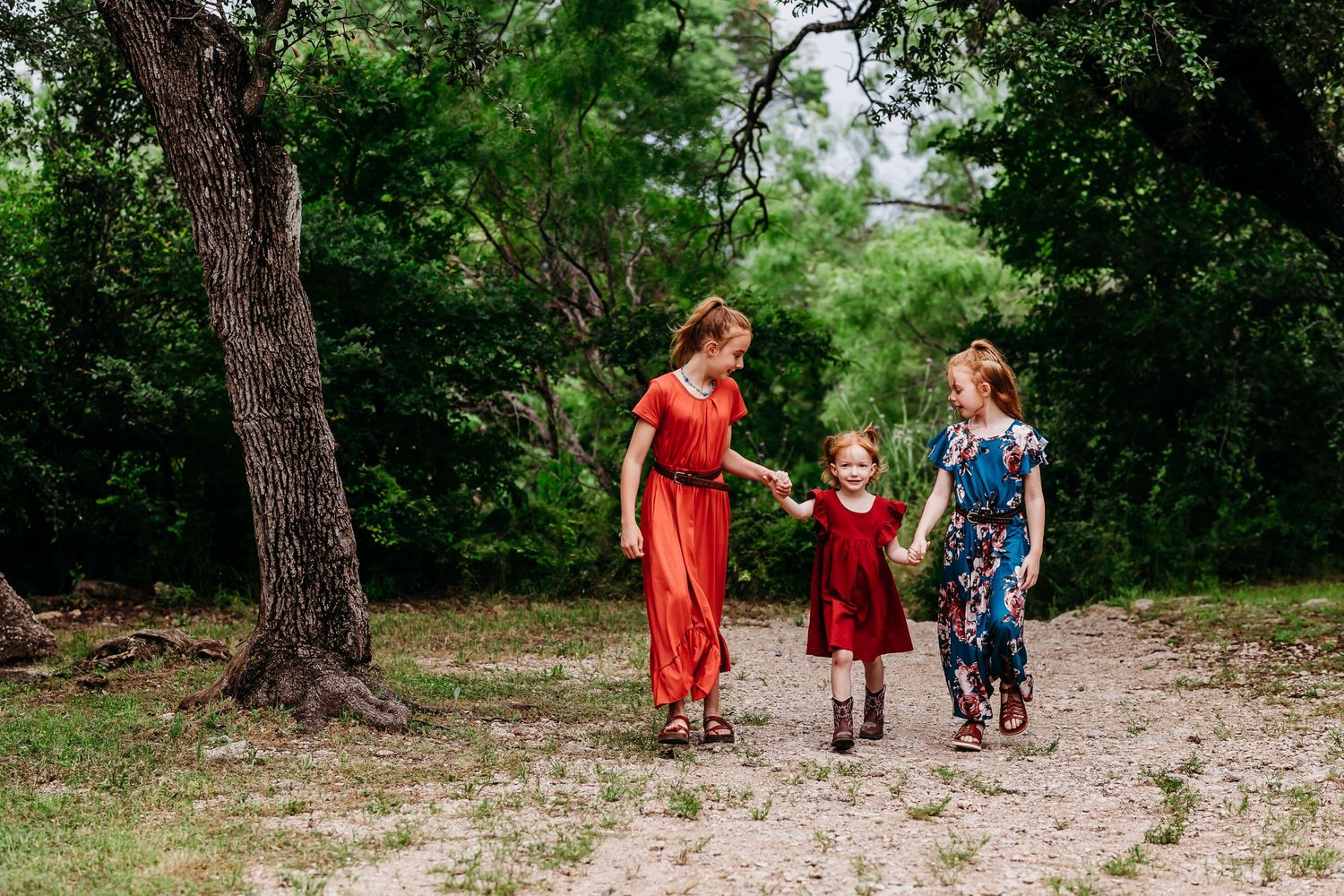Three people walking together on a dirt path through a wooded area wearing colorful summer dresses in red and blue.