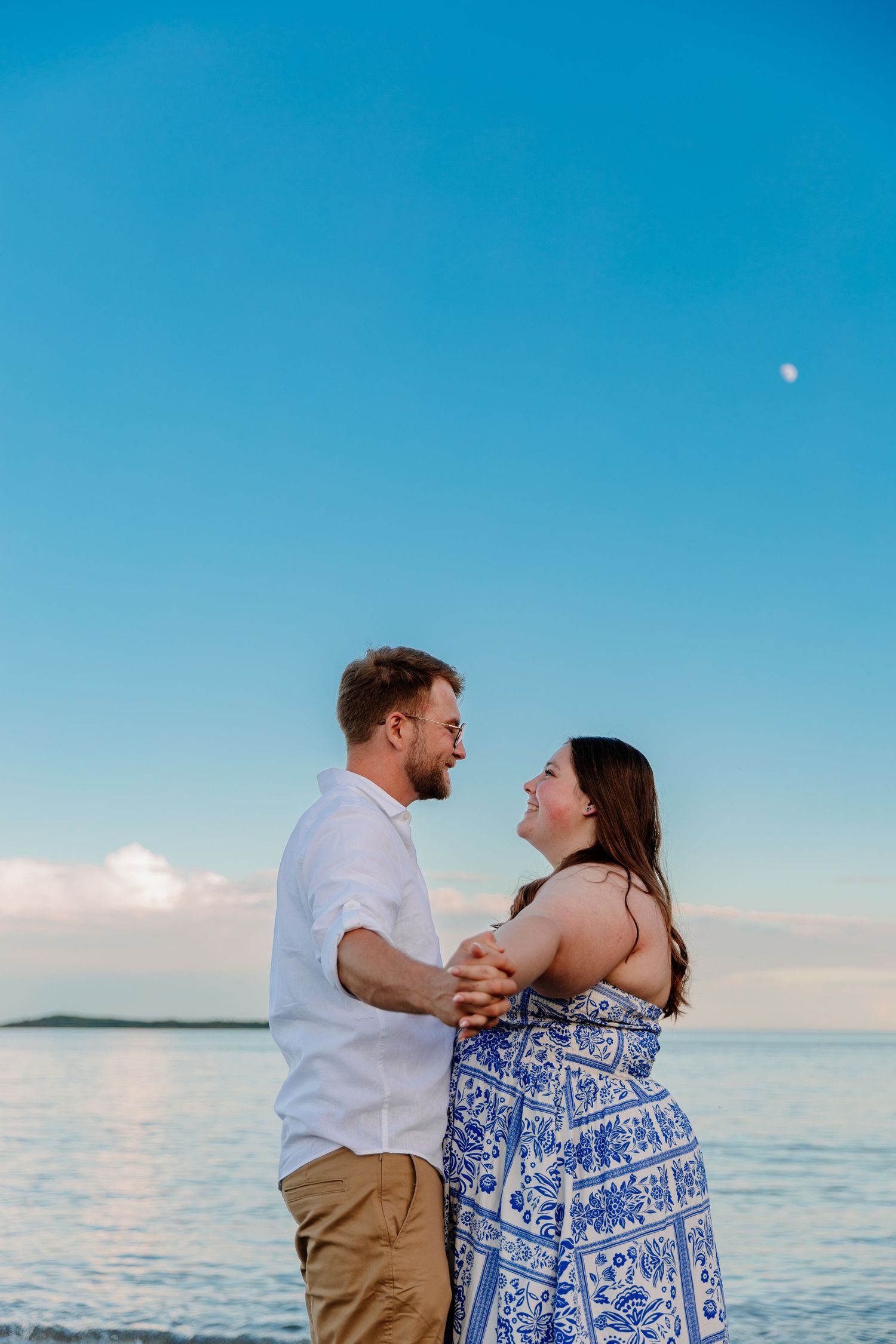 Couple holding hands and smiling at each other on a beach at sunset with blue sky and ocean backdrop.