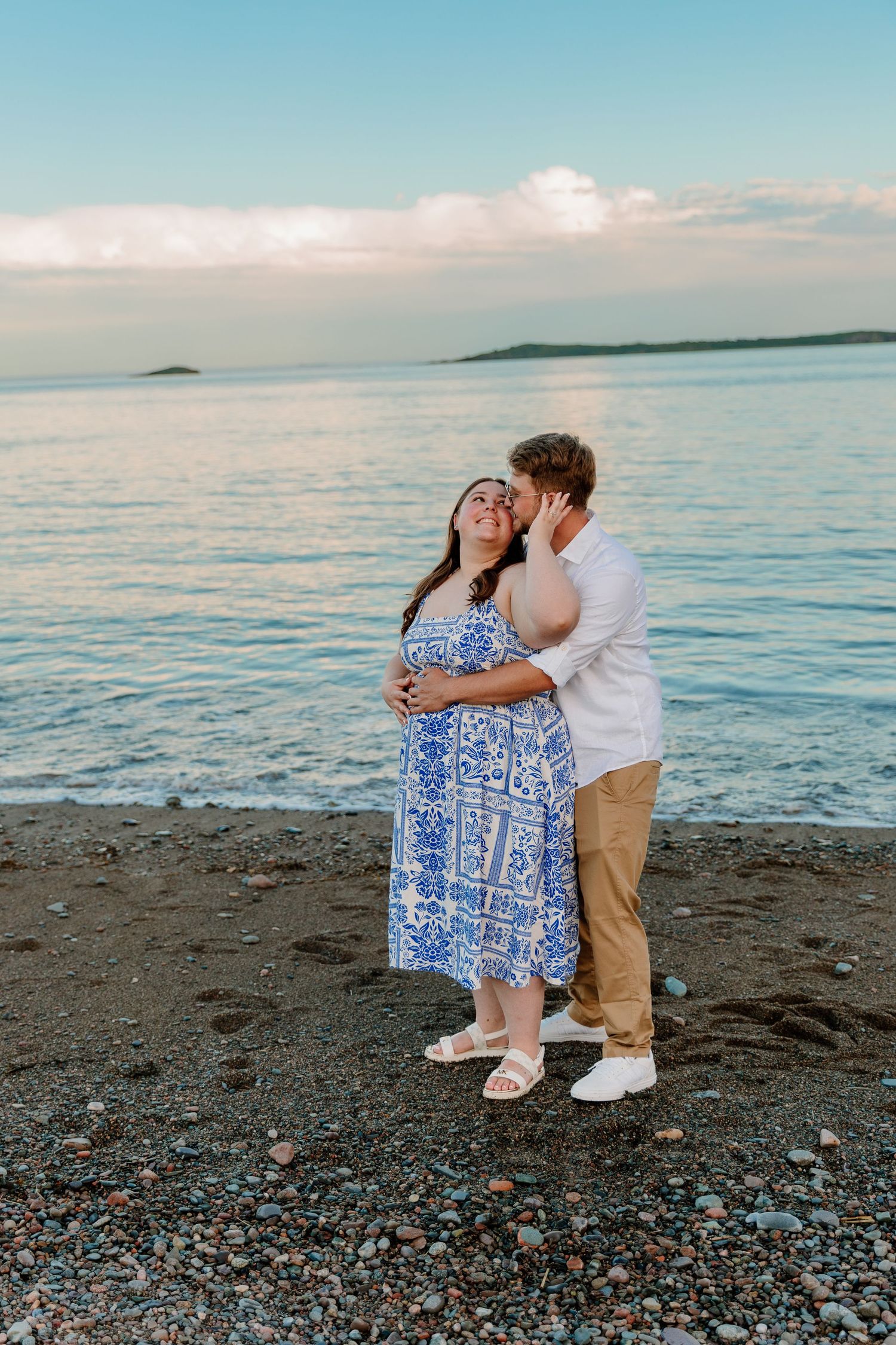 Couple sharing a romantic kiss on a rocky beach at sunset, woman in blue floral dress cradling baby bump.