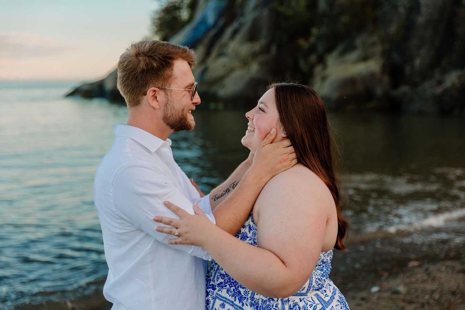 Couple sharing a romantic moment by the water at sunset, man holding woman's face as they smile at each other.