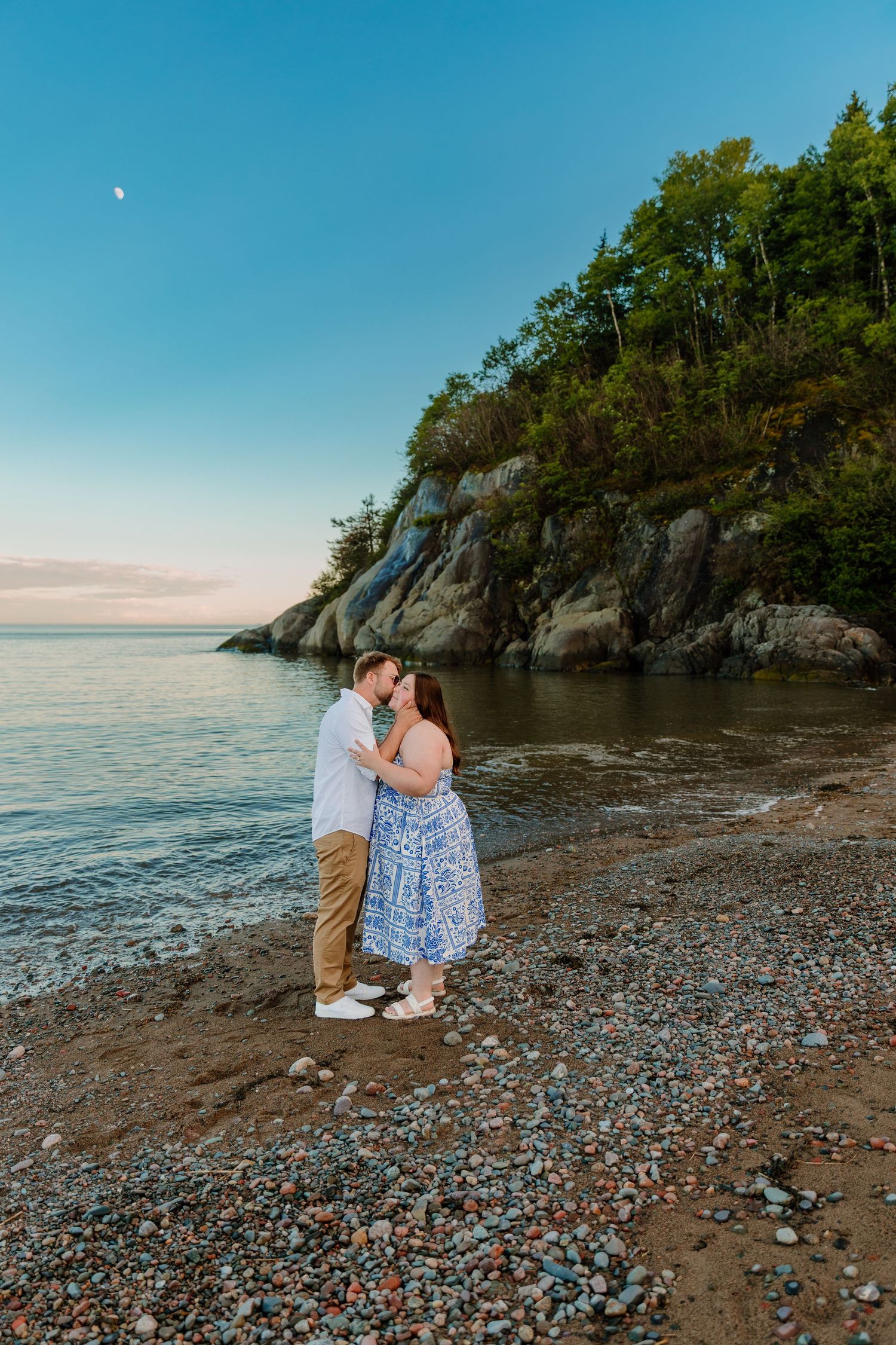Couple kissing on a rocky beach at sunset with lush cliffs and calm water in the background.