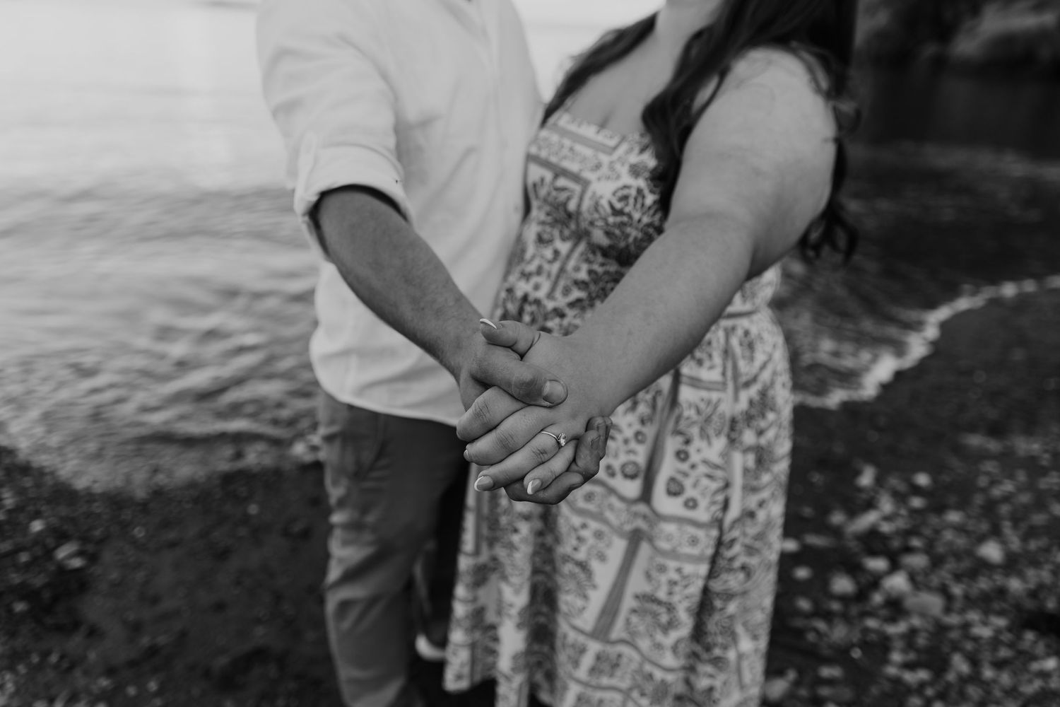 Black and white photo of a couple holding hands on a rocky beach shore, woman in floral dress.