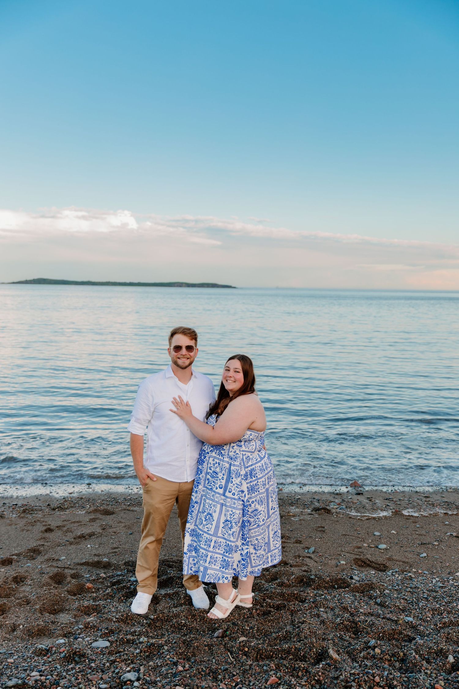 Couple smiling together on a rocky beach at sunset, woman in blue floral dress, man in white shirt and sunglasses.