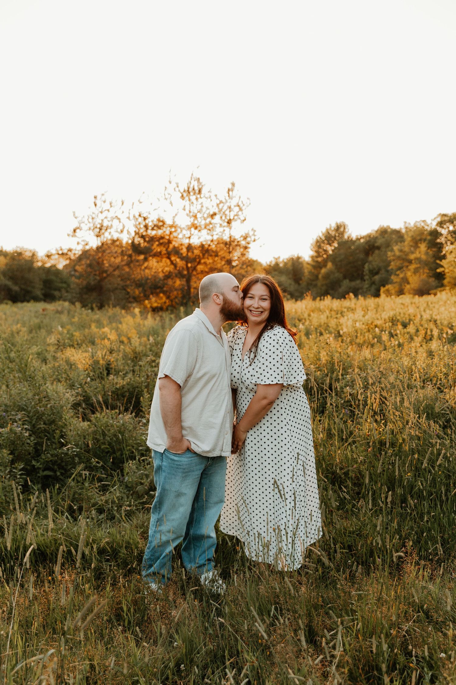A couple shares a tender kiss in a golden sunlit meadow, the woman in a floral dress smiling joyfully.