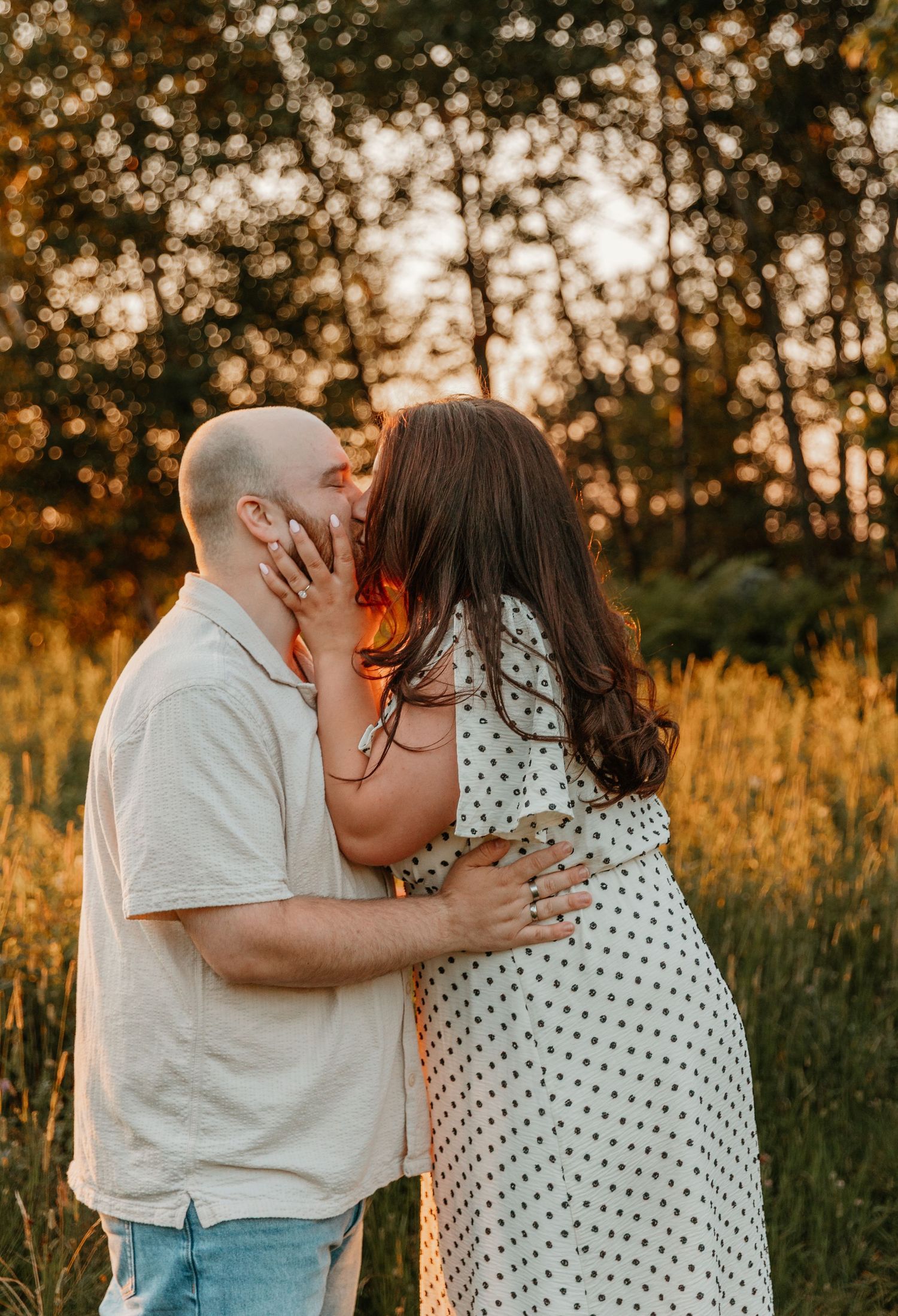 Couple sharing a romantic kiss at golden hour in a sunlit meadow, woman in polka dot dress embracing bald man.