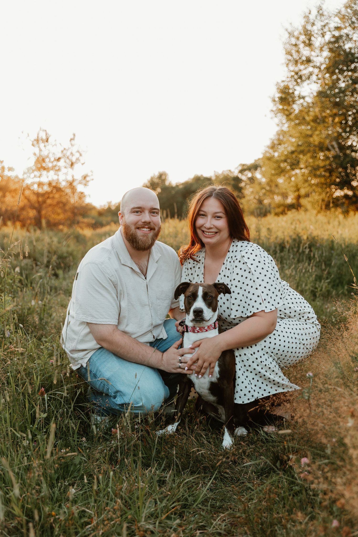 A smiling couple crouches in a sunny field with their black and white dog, surrounded by lush green grass and trees.