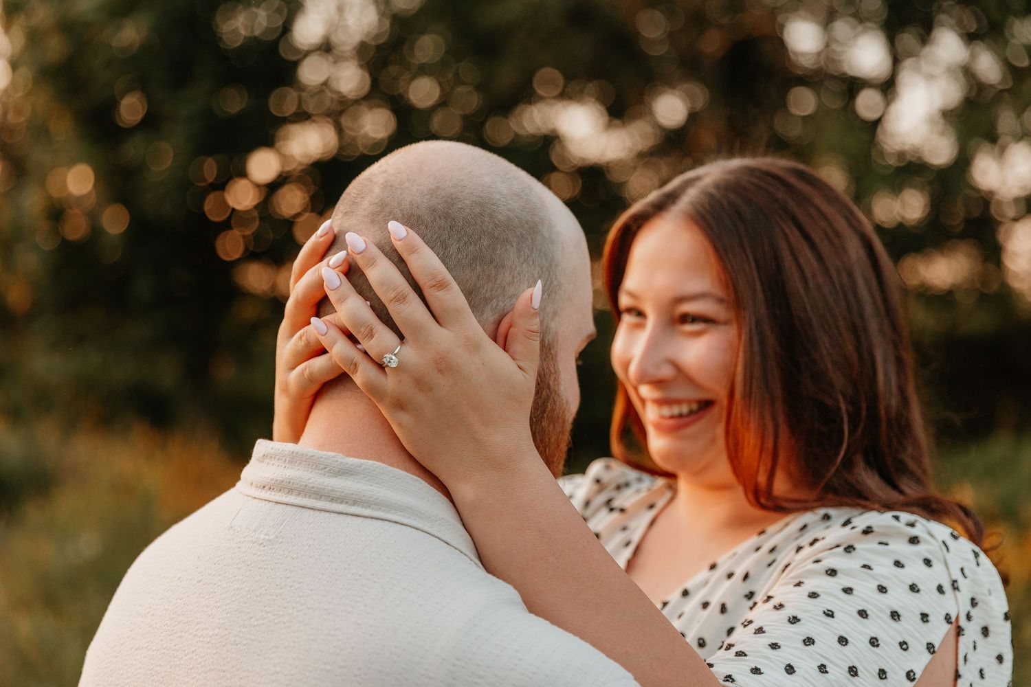 Smiling woman with engagement ring embracing bald man outdoors in golden hour light.