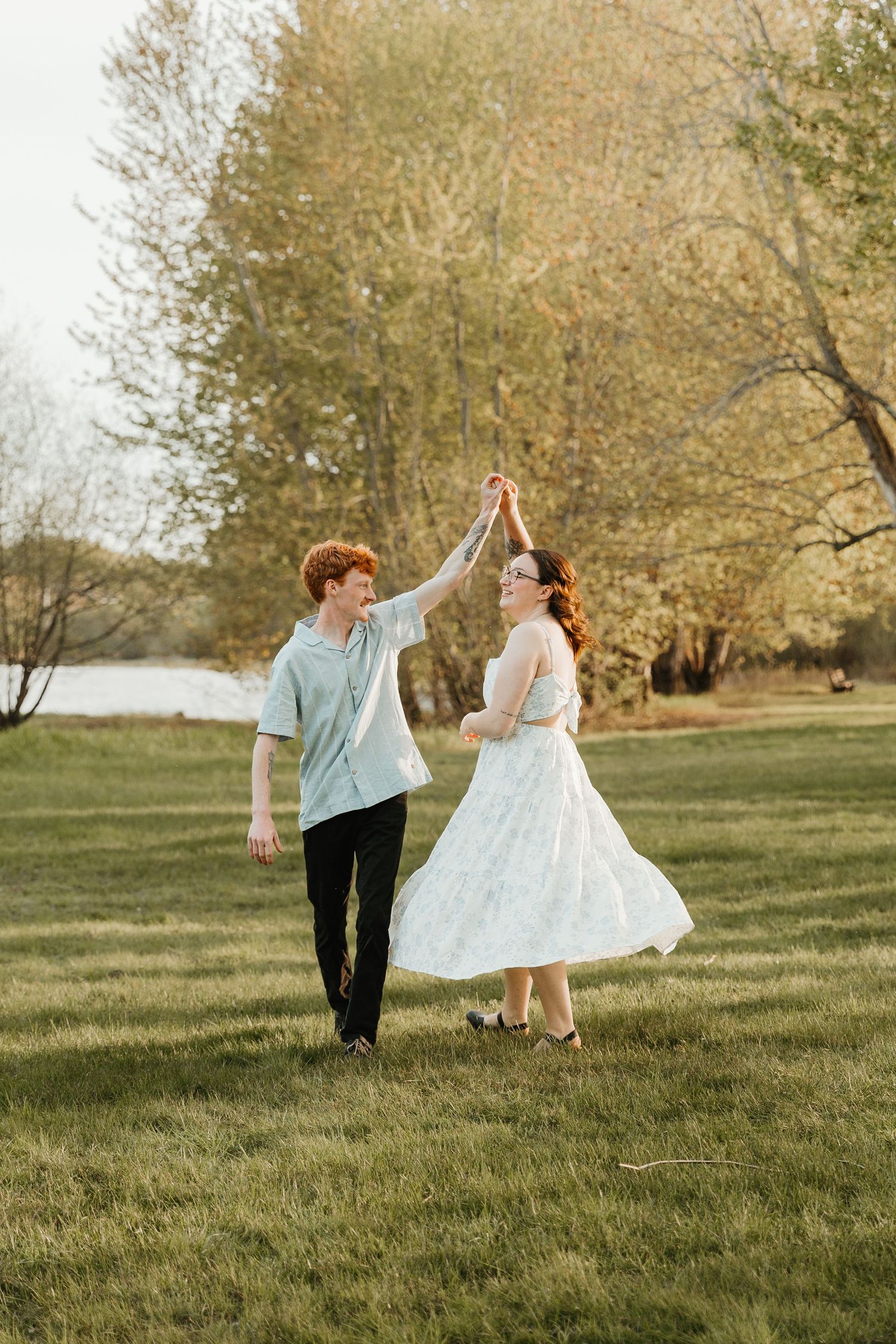 A couple dances outdoors on green grass, woman in white dress, man in blue shirt, surrounded by spring trees.