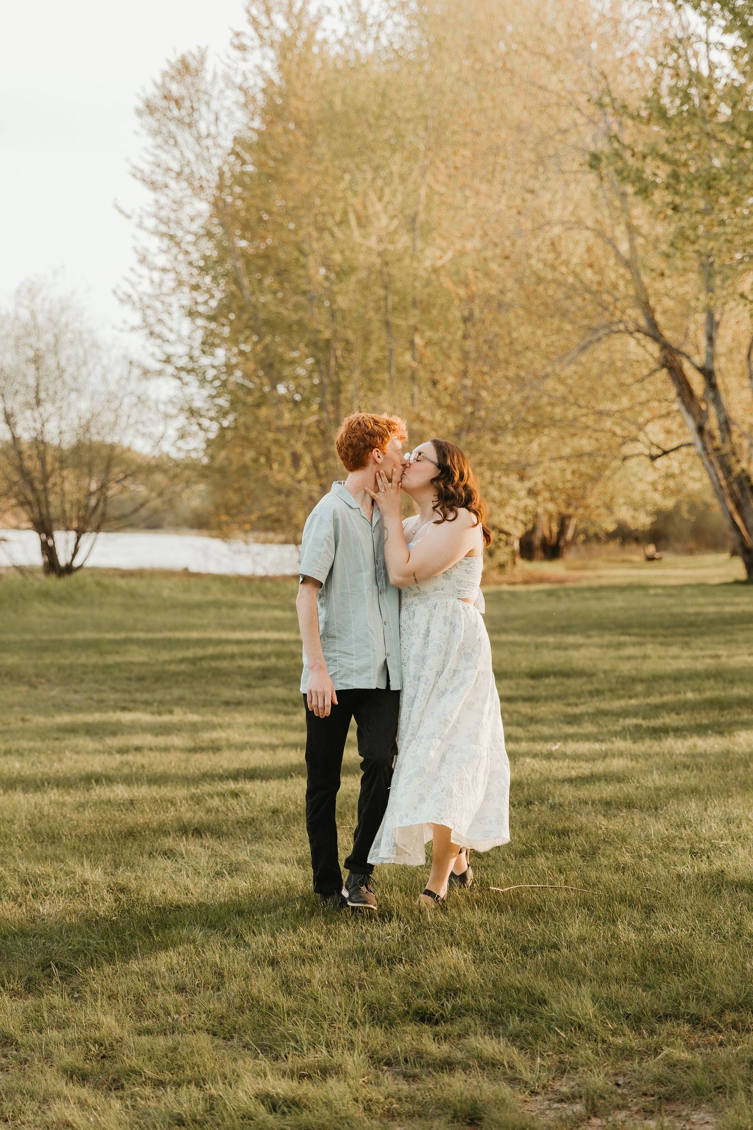 A couple shares a romantic kiss in a sunlit park, surrounded by lush green grass and golden trees near a lake.