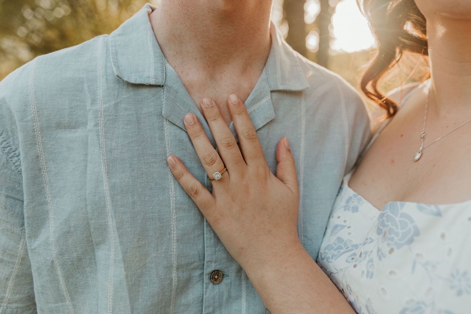 Close-up of a woman's hand with an engagement ring resting on a man's chest outdoors.