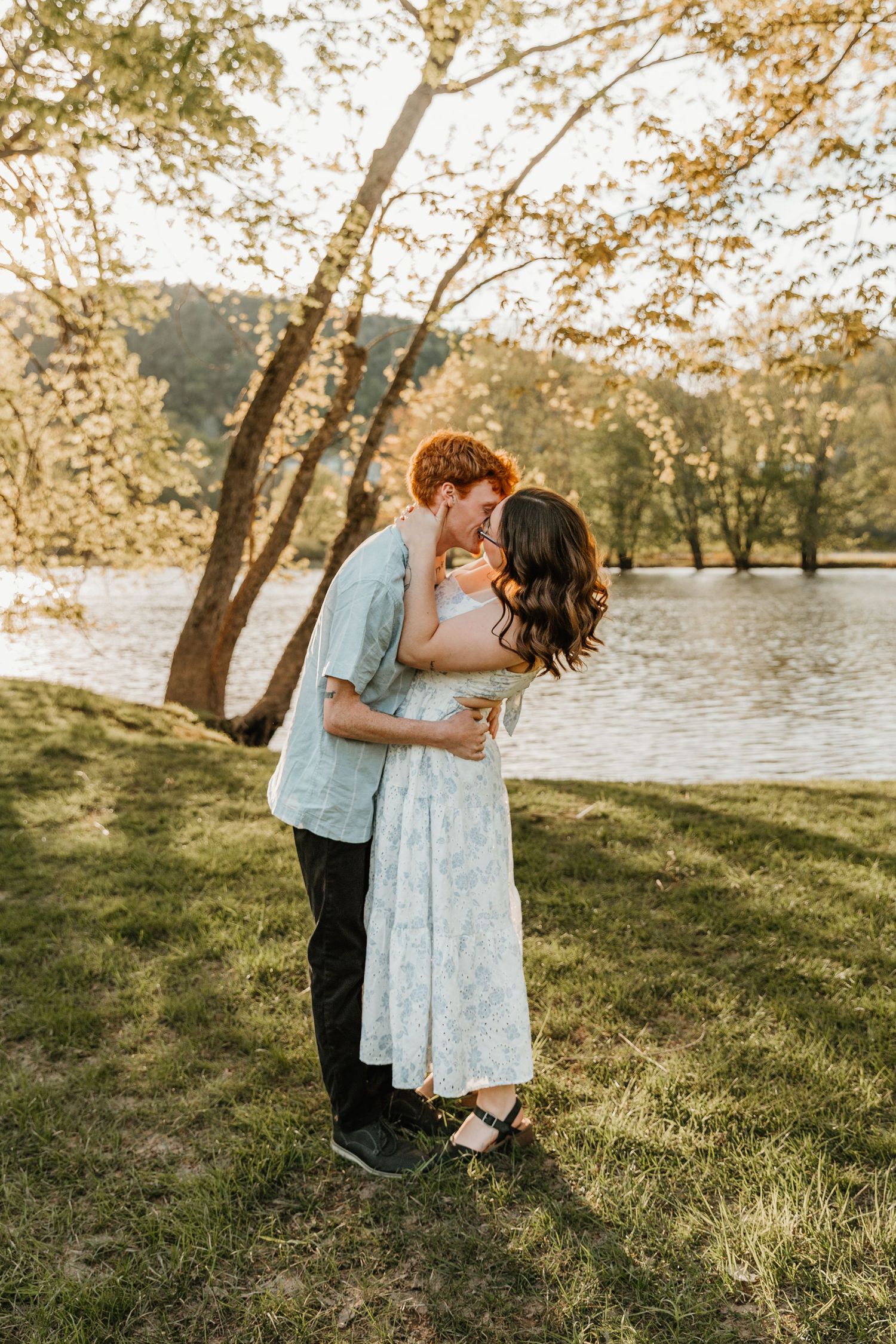 A couple shares a romantic kiss by a sunlit lake, surrounded by lush green trees, the woman in a floral dress.