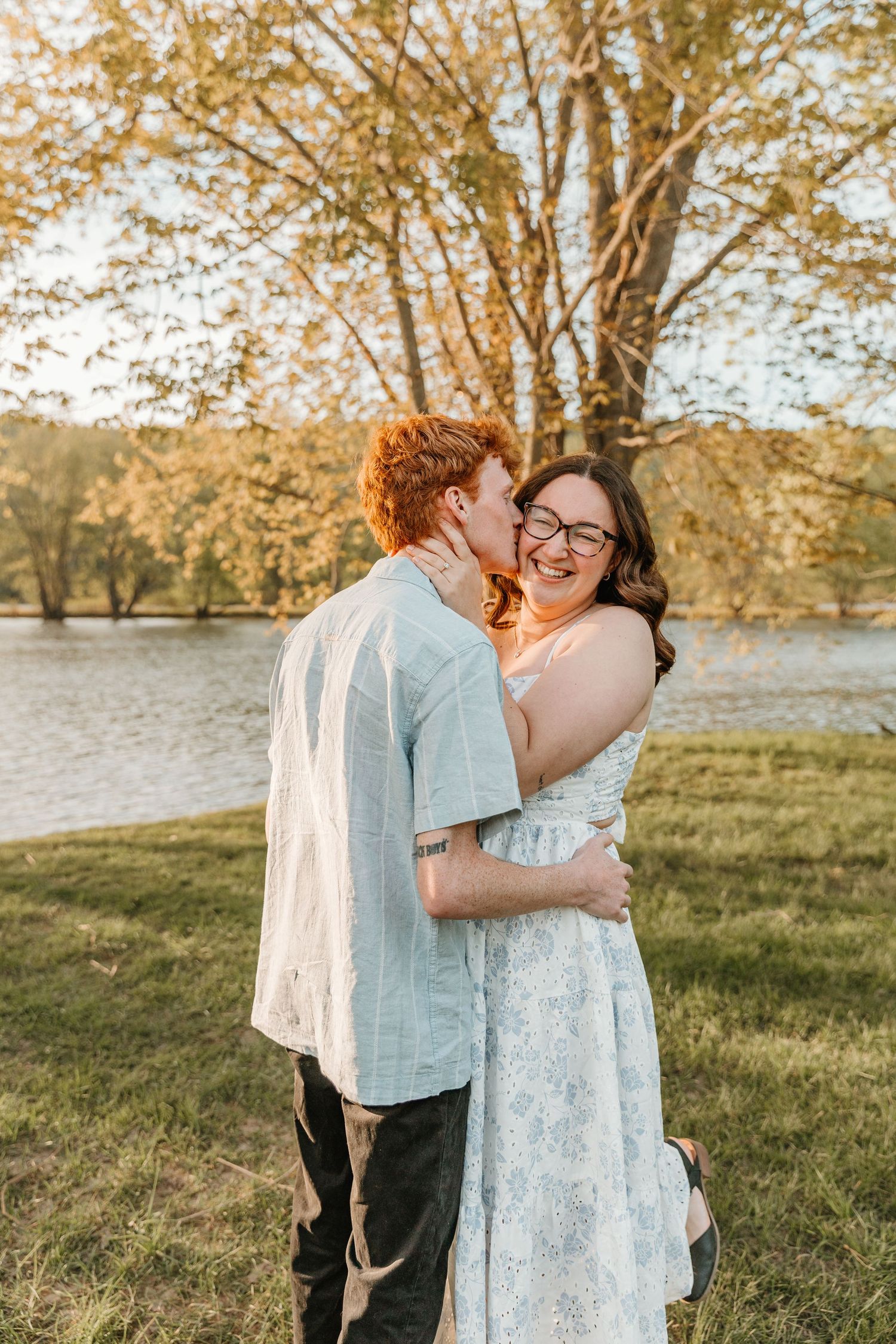 A couple shares a tender kiss outdoors near a lake, surrounded by golden autumn trees at sunset.
