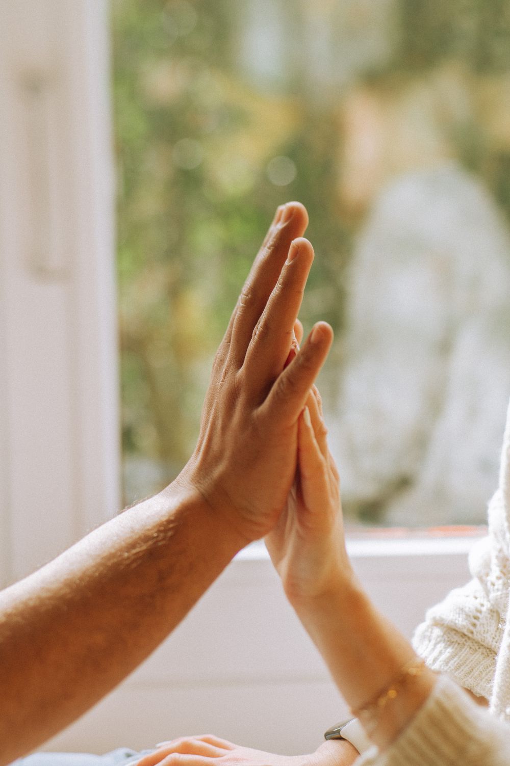 Two hands reaching towards each other against bright window background.