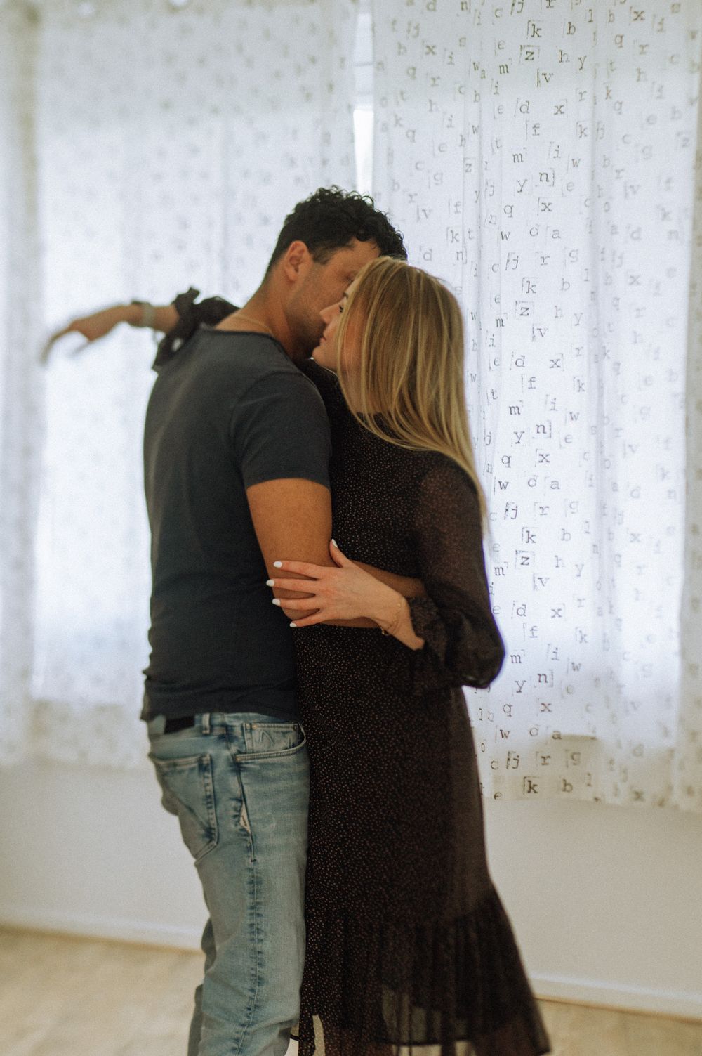Couple embracing in bright white room with patterned curtains.