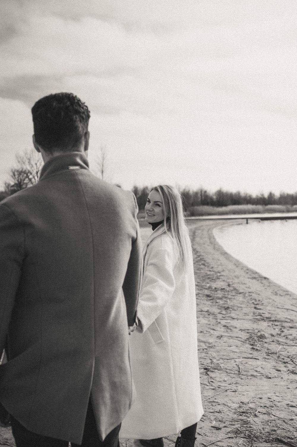 A black and white photo of a couple walking along a winter shoreline.