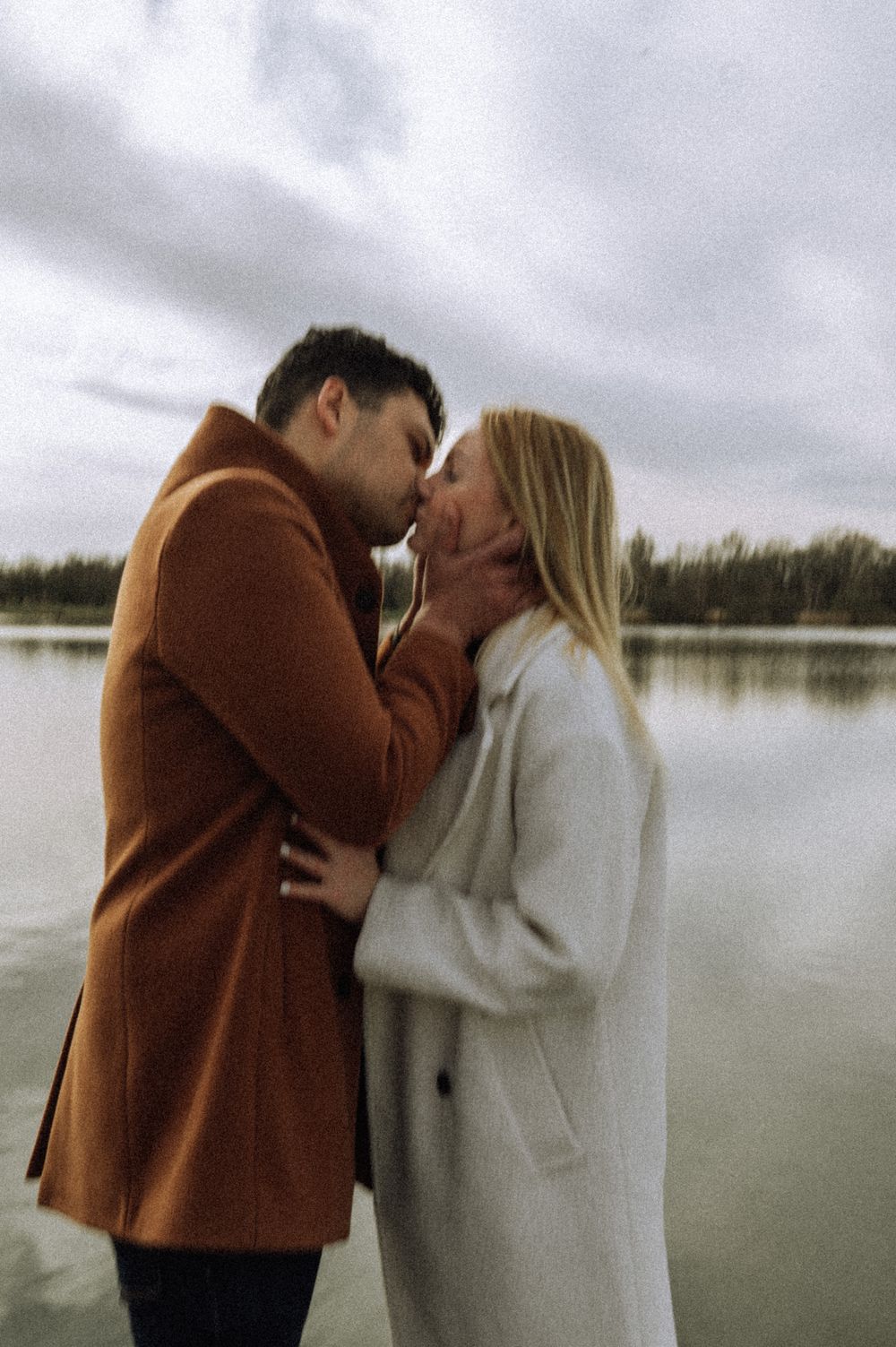 Romantic couple embraces by a serene lake on an overcast day, wearing brown and white coats.
