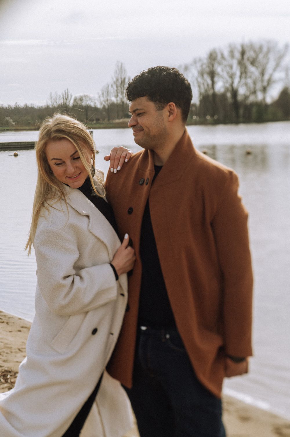 Intimate couple shares tender moments on a beach wearing contrasting brown and white outerwear.