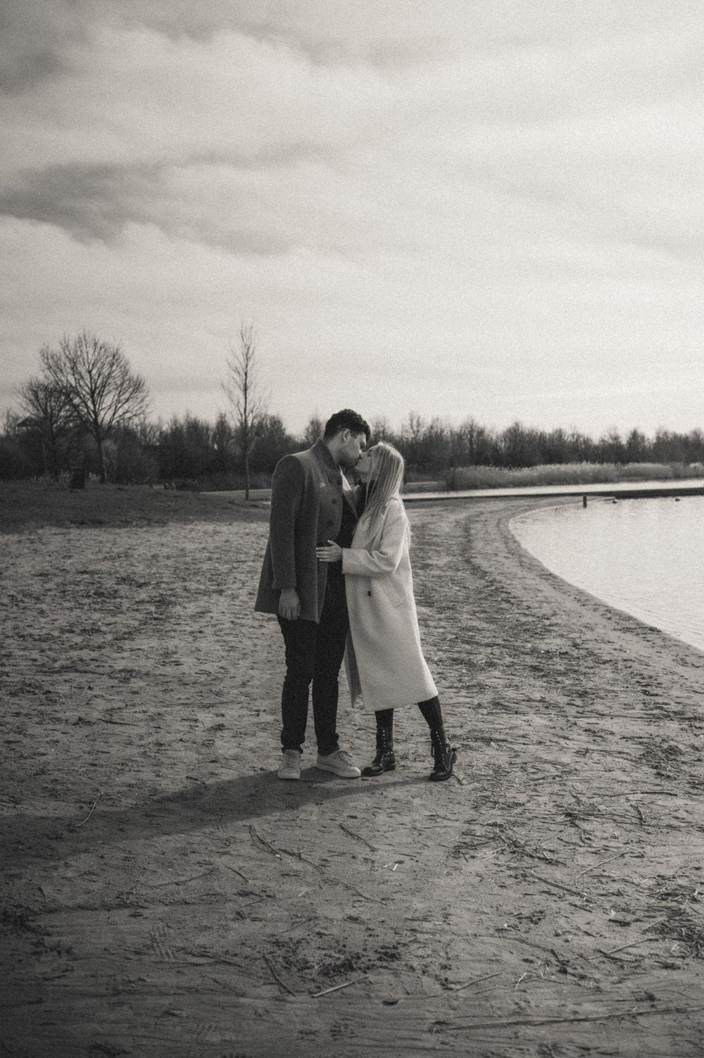 Couple sharing intimate moment on sandy beach with water and trees in background.