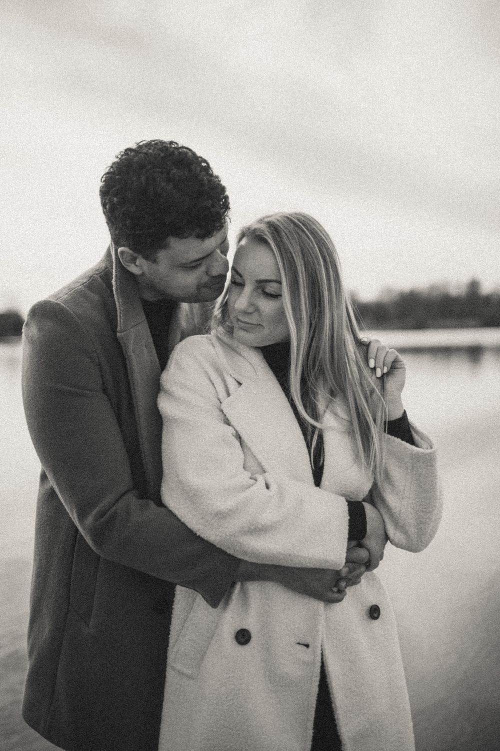 Romantic black and white photos of couple in winter coats by waterfront.