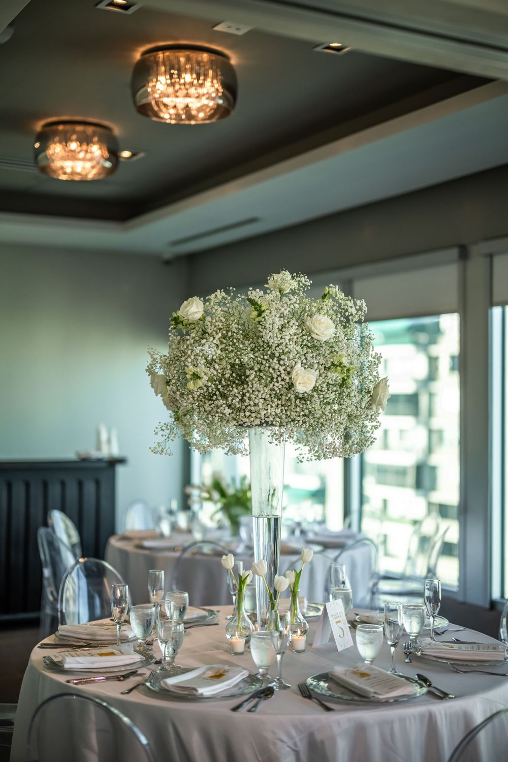 Tall baby's breath centerpiece on round reception table under crystal chandeliers.