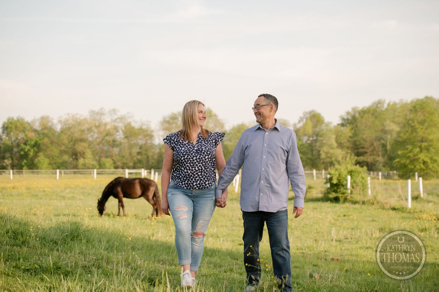 A couple walks together through a green pasture with horses and fencing in the background.